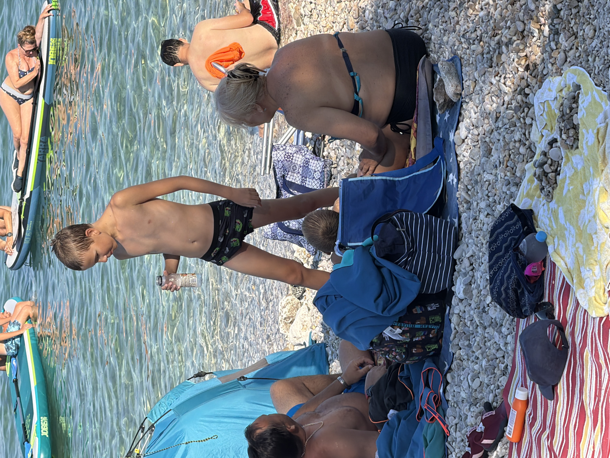 This is a vertical, eye-level shot of a sunny day at a pebbled beach in Općina Baška, Croatia. The water is clear and shallow, with sunlight reflecting off its surface. In the foreground, a young boy, wearing patterned swim trunks, holds a clear plastic bottle and looks down. Next to him, a man with a silver chain around his neck lies on a blue towel under a light blue beach tent. Scattered around them are various beach items, including towels (one yellow and white striped, another red and white striped), bags, bottles, and a dark baseball cap.
In the background, several people are enjoying the beach. A woman in a black and white polka dot bikini is on a paddleboard in the water. Behind her, a man with fair skin, wearing red and black swim trunks, is partially visible. Another woman, with blonde hair and wearing a dark bikini, is seated on the pebbles, her back to the camera. The scene suggests a relaxed family outing on a warm summer day. The water's clarity and the abundant sunshine indicate pleasant weather.