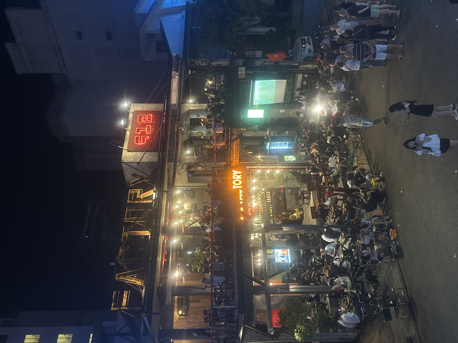 The image presents a vibrant nighttime urban scene, centered on a multi-story establishment identified by prominent illuminated signs reading "FACTORY" and "612 BISTRO BEER." This location is situated in Thuận Hóa Ward, Huế, Vietnam. The building features an open-air industrial aesthetic with exposed metal framework and multiple levels for patrons.
On the building's upper balcony level and the first-floor balcony, numerous individuals are seated at tables, engaged in socializing and dining or drinking under string lights and spotlights. The ground level of the establishment also hosts patrons seated at tables within a semi-open area.
Extending from the ground floor onto the asphalt street, a significant crowd of dozens of people, predominantly adults, are gathered. Many are seated at small tables and stools directly on the street, while others stand and interact. To the right of the ground floor entrance, a large white projection screen displays an image, likely a video or live event, which a segment of the street crowd is observing. A Vietnamese flag is mounted to the right of the projector screen. A white sedan is parked on the street further to the right. The entire scene is brightly illuminated by various artificial lights, contrasting with the dark night sky and dimly lit surrounding buildings in the background.