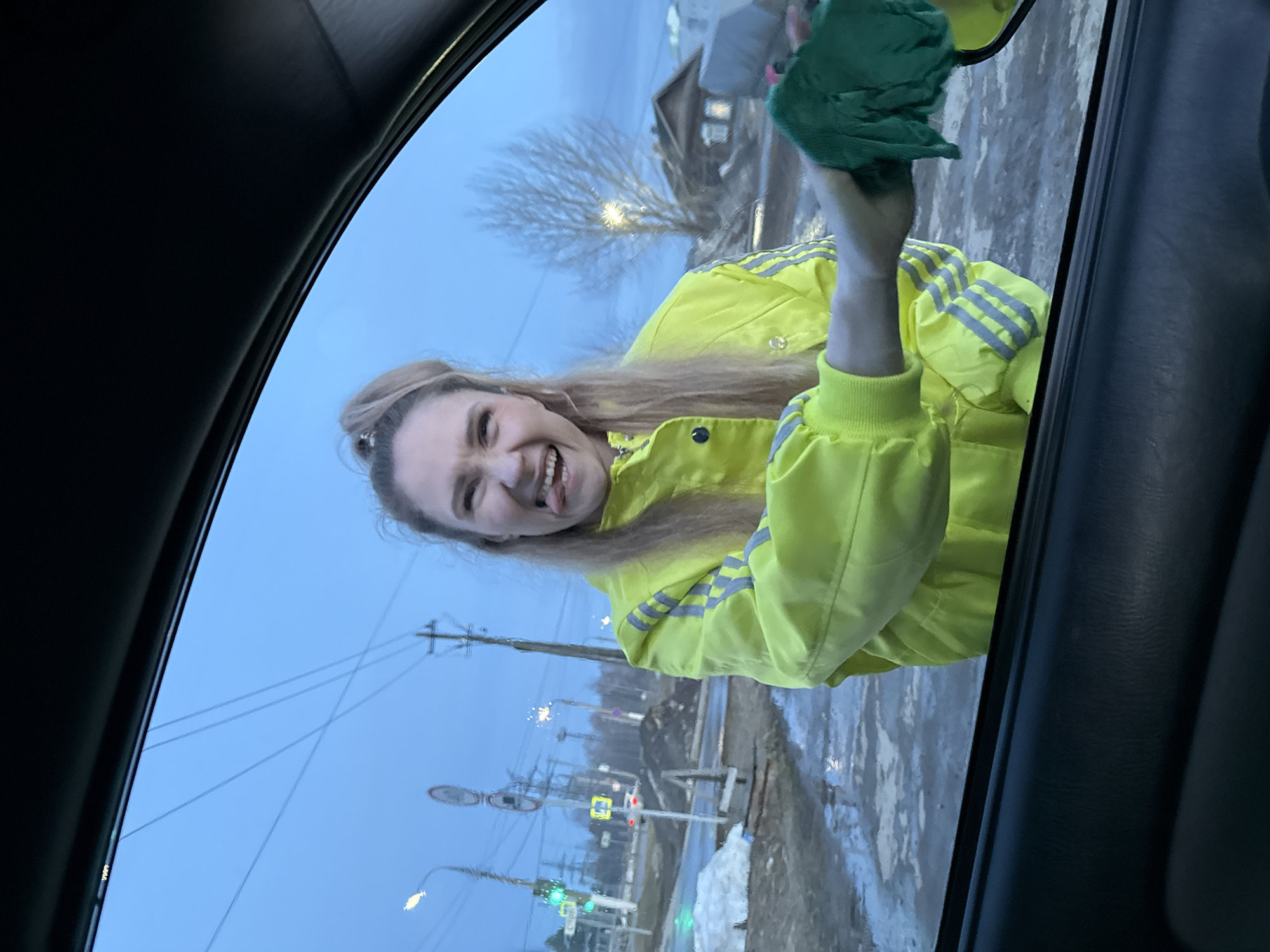 The image captures a scene from inside a vehicle, looking out the driver's side window, in the city of Chaikovsky, Russia. A young adult female, with light-colored hair tied in a high ponytail, stands outside the vehicle, facing the camera. She is wearing a bright neon yellow jacket with reflective silver stripes on the sleeves. Her facial expression is animated, featuring a wide, open-mouthed smile with her tongue playfully extended. Her right arm is extended towards the right side of the frame, holding a crumpled dark green cloth, which she is using to wipe the vehicle's side mirror, partially visible.
The background depicts an urban street environment under a dull, overcast sky, indicative of dusk or early morning. The ground is wet and irregular, with patches of dirty snow and slush visible on the left side near the street. Visible street furniture includes a street light pole, a traffic light showing a green signal, a pedestrian crossing sign, and a circular red and white road sign. Utility poles with multiple overhead wires crisscross the sky. In the mid-distance, a bare deciduous tree stands, and a traditional wooden house with glowing windows is discernible to the right, suggesting residential architecture. The overall scene suggests a transitional season, likely late winter or early spring.