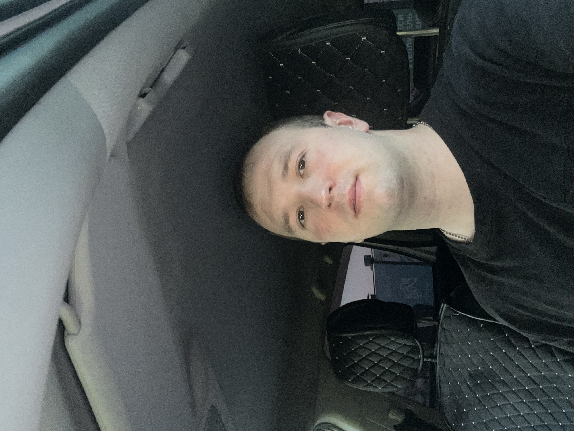 The image shows a man inside a car. He is wearing a black t-shirt and a silver necklace. The car's interior features a quilted pattern on the seats. Outside the car's window, there's a partial view of a building with what appears to be Cyrillic text. Based on the provided information, the location is Chaikovsky, Russia.