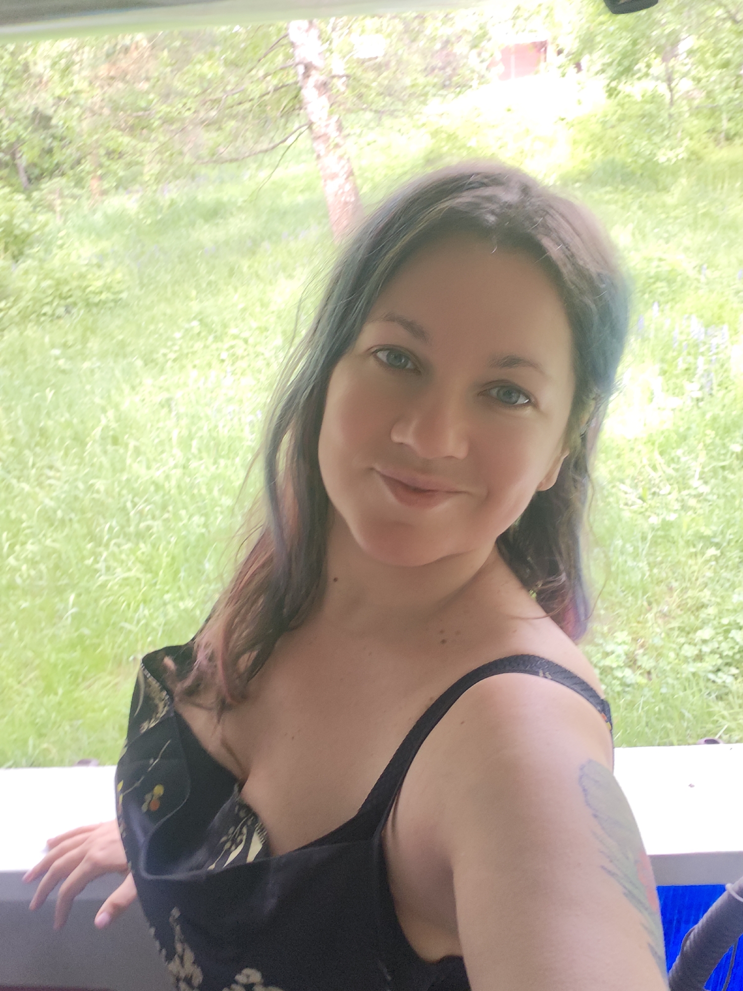 A woman is posing for a selfie. She has fair skin, blue eyes, and dark hair with blue and pink highlights. She is wearing a black dress with a floral pattern and black spaghetti straps. Her right arm is visible with a tattoo on her upper arm. She is standing on a white porch or balcony overlooking a green grassy area with trees. The background is blurred, suggesting depth of field. The location is possibly Свистуха (кузяевский С/о), Russia, although this cannot be confirmed from the image alone.