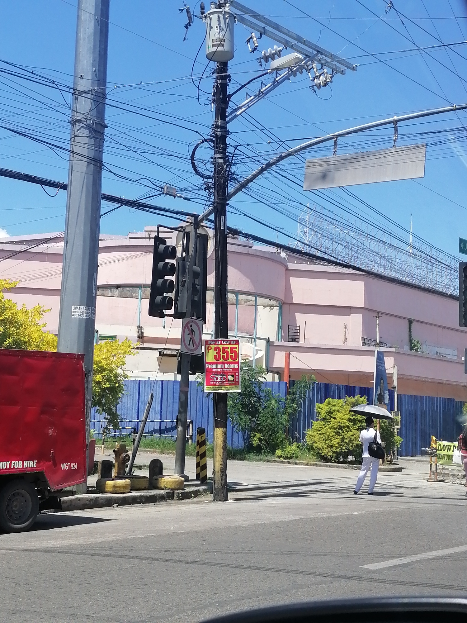 The image depicts a street scene in Davao City, Philippines, with a focus on a traffic intersection. A prominent traffic light is visible, with the red and yellow lights illuminated. To the left of the traffic light, a red truck with the text "NOT FOR HIRE WGT 924" is parked near a fire hydrant and several tires that appear to be used as barricades.
In the background, a large, pink building with arched windows and a patterned facade stands out. Blue construction barriers are erected in front of the building, and several trees with vibrant green foliage are present. A person dressed in white, carrying an umbrella and a dark bag, is crossing the street. Another person, partially visible, is also crossing the intersection.
Overhead, a complex network of electrical wires and a utility pole with a transformer are visible against a clear blue sky. A blank rectangular sign hangs from a cable above the intersection, and a smaller sign advertising "Pay as low as P355 Premium Rooms" is attached to the traffic light pole. The overall scene suggests a typical urban environment in Davao City.