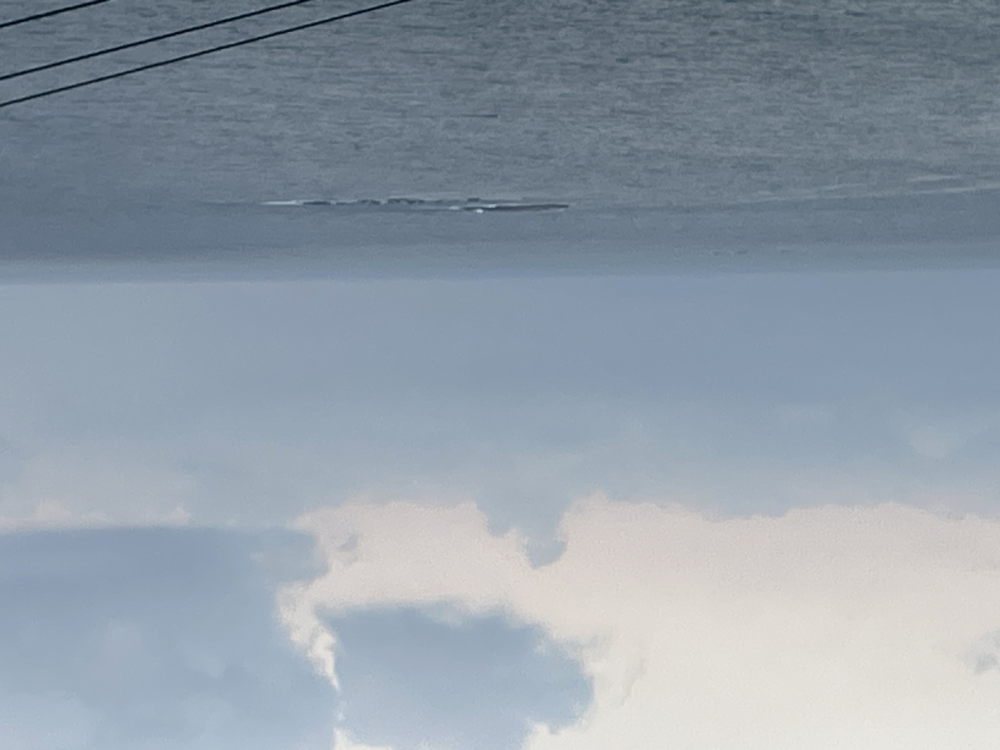 The image captures a wide view of the sea under an overcast sky. In the middle distance, a long, low-lying island or rock formation is visible in the water. Waves are breaking around the formation. The sky is filled with soft, muted clouds in shades of white and pale blue. The sea itself is a deep grey-blue, with subtle ripples on its surface. In the foreground, three dark, diagonal lines, likely power lines or cables, run across the bottom of the frame. The overall impression is one of a calm, serene, and somewhat melancholic seascape. The location of Mate Asher Regional Council in Israel is a coastal region in northern Israel, known for its scenic beauty and proximity to the Mediterranean Sea. This image is consistent with such a landscape.