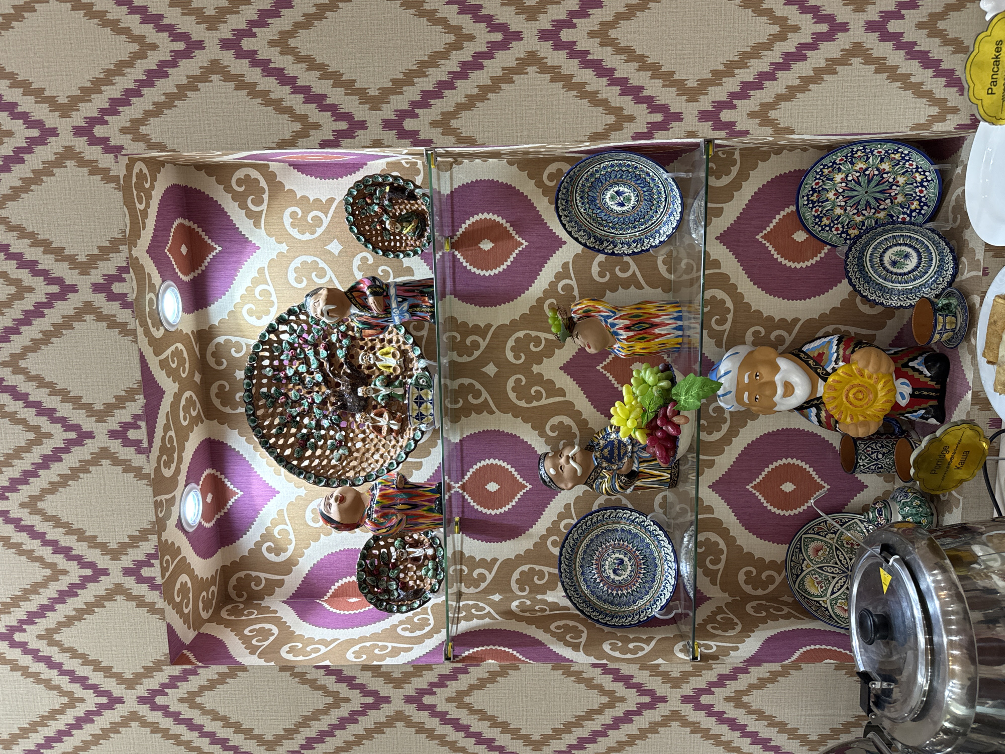 The image shows a display shelf or counter in a room, likely in Samarkand City, Uzbekistan, given the presence of traditional Uzbek ceramics and figures. The background wall features a repeating geometric pattern in shades of beige and purple. The counter or shelf is lined with a patterned fabric featuring large, stylized motifs in beige, brown, and purple.
On the shelf, there are several decorative items, including:
* Hand-painted ceramic plates of various sizes, featuring intricate floral and geometric patterns.
* Figurines of men dressed in traditional Uzbek clothing.
* Decorative bowls filled with colorful objects.
* A metal food warmer or container.
* A sign labeled "Pancakes".
The shelf is divided into sections by vertical glass panels. The overall impression is of a display of Uzbek cultural artifacts, possibly in a shop, museum, or hotel setting. The items are arranged with attention to aesthetics.