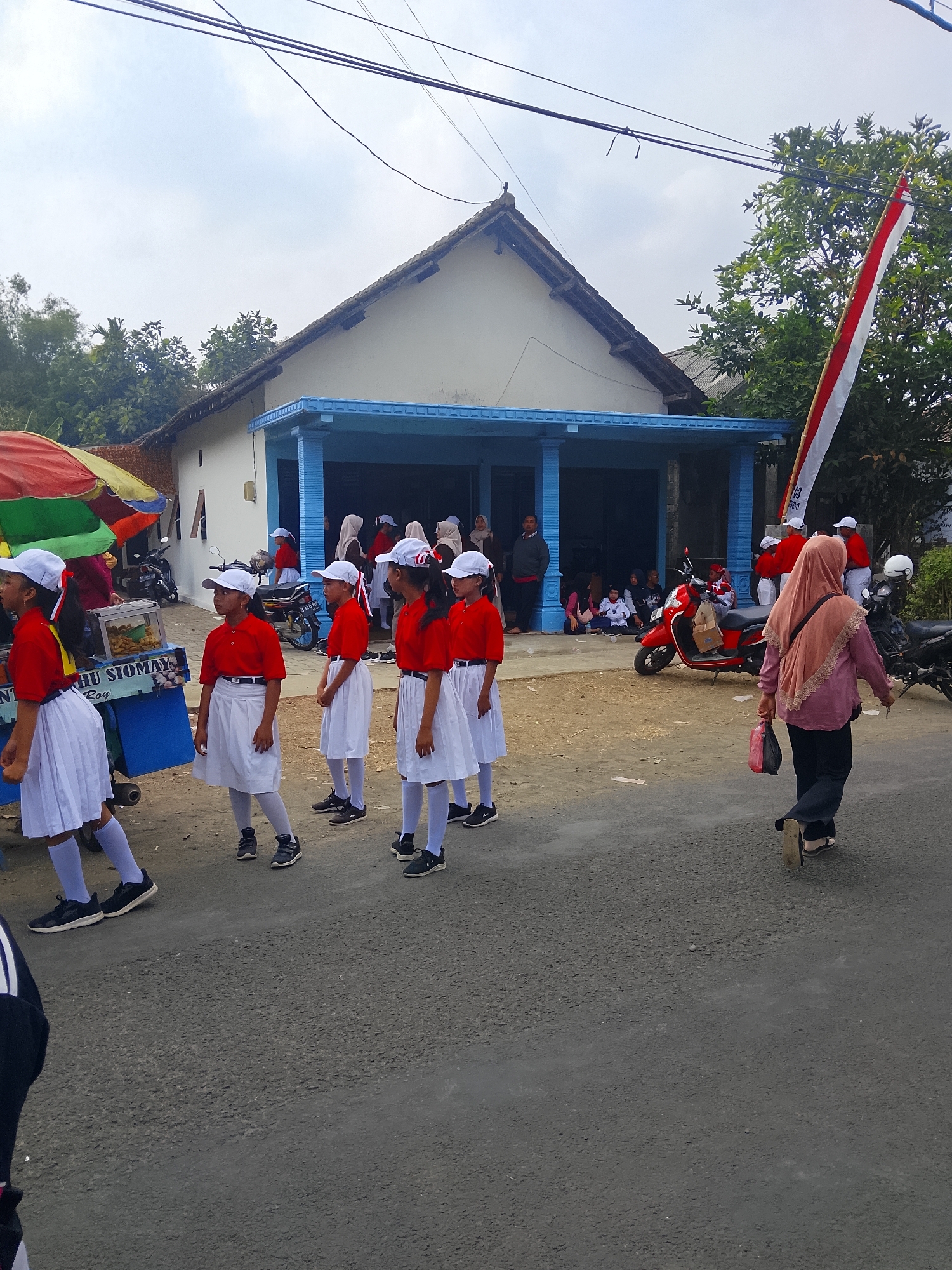 In Bangsri, Indonesia, a group of young girls in red shirts and white skirts, wearing white caps, stand in a line on a paved road. They appear to be participating in a parade or event, as evidenced by their matching uniforms and the Indonesian flag pole visible in the background. Behind them, a building with a blue-covered entrance and white walls stands. Several motorcycles are parked near the building, and a few adults and children are gathered outside. A food cart with a colorful umbrella is also present on the left side of the image, with text on its side that reads "SIOMAY". A woman in a pink hijab and dark pants walks away from the camera, carrying two plastic bags. The sky is overcast, and trees are visible in the distance.