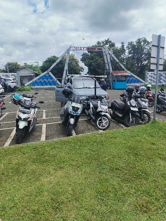 The scene is located at the "Gerbang Wisata Alam" (Nature Tourism Gate) in Majalengka, Indonesia, specifically at the coordinates -6.846 latitude and 108.204 longitude. The image depicts a paved parking area in front of a metal archway with a sign that reads "Gilava Wisata Alam" and a blue, white, and grey patterned backdrop. The parking area is partially covered by a grass lawn. There are five motorcycles parked in a line, facing the archway. Two of the motorcycles are white and black, one is blue, and the other two are black. There is a grey van parked behind the motorcycles, partially obscured by the motorcycles, with a driver and a passenger in the front seats. Behind the van, there are two people visible on the right side of the image, one sitting on a blue bench. The people appear to be casually observing the area. There are several cars parked in the background. The sky is partly cloudy, and the air is clear.