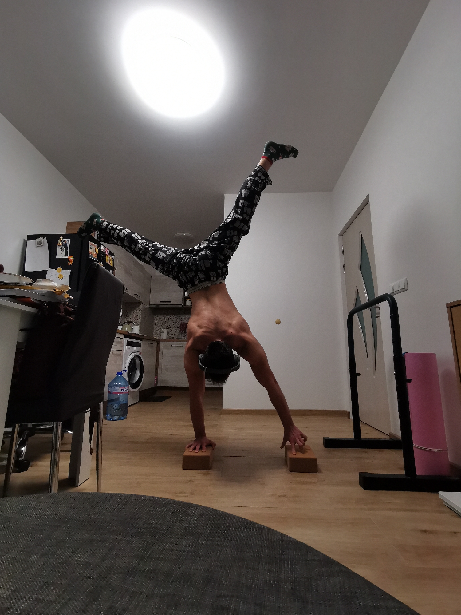 A person is performing a handstand on yoga blocks in an indoor setting. The person is shirtless and wearing patterned pants and socks. One leg is pointing upwards, while the other is angled out to the side. The person appears to be in a room with a kitchen area visible in the background, including a washing machine and cabinets. There's also a desk with a chair and a water bottle in the foreground. A door with a pull-up bar and a pink yoga mat are visible on the right side of the image. The lighting is bright, coming from an overhead light. There is no evidence to suggest this is occurring in the city of Burgas, Bulgaria.