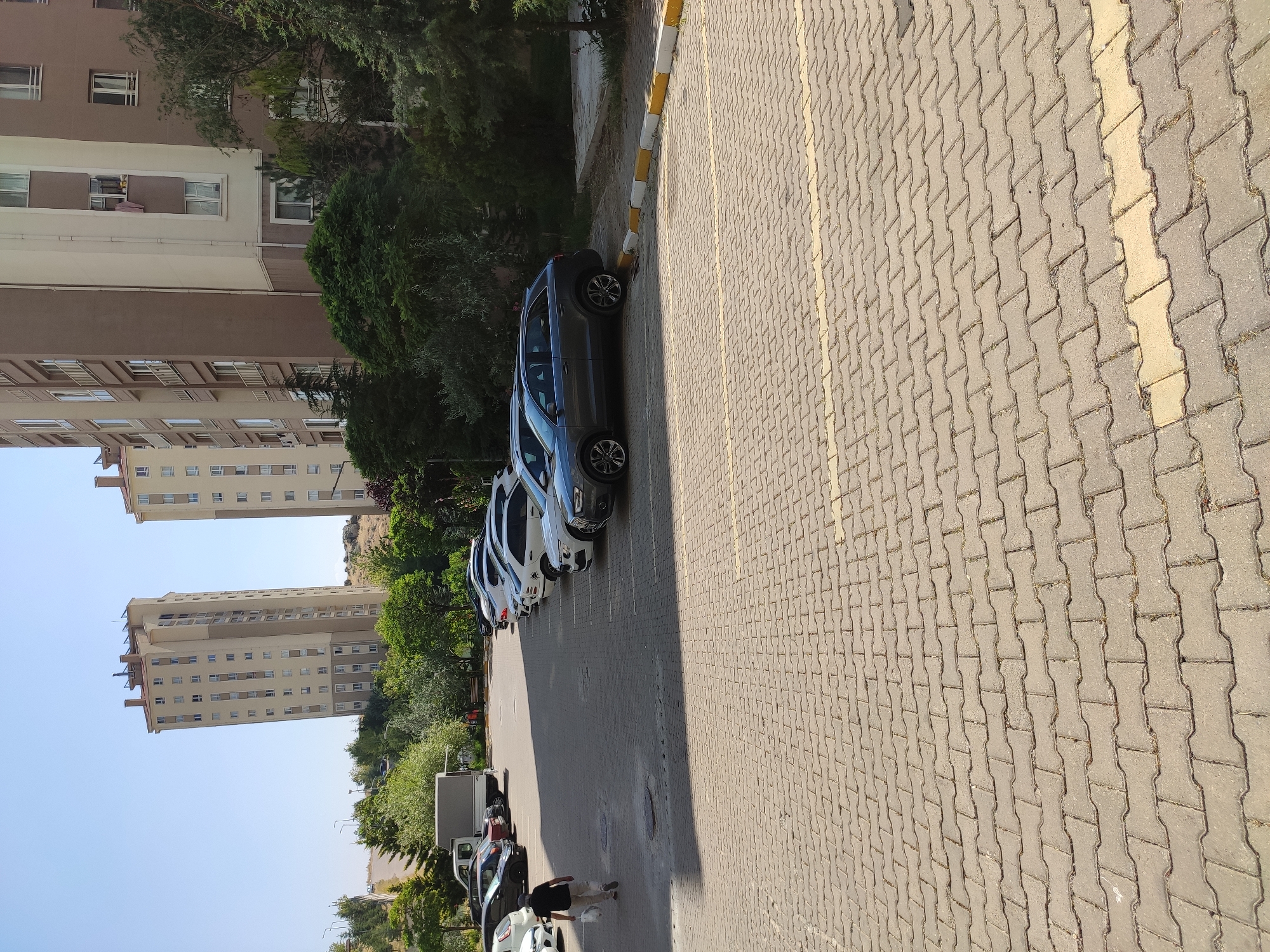 A man in a dark t-shirt and light-colored pants walks away from the camera across a paved parking lot in Etimesgut, Turkey. He is carrying a white bag in his left hand. The parking lot is composed of interlocking concrete pavers and is marked with yellow lines. In the background, a row of parked cars, including a dark gray SUV, is visible. Behind the cars are several apartment buildings and trees. The sky is clear and blue.
