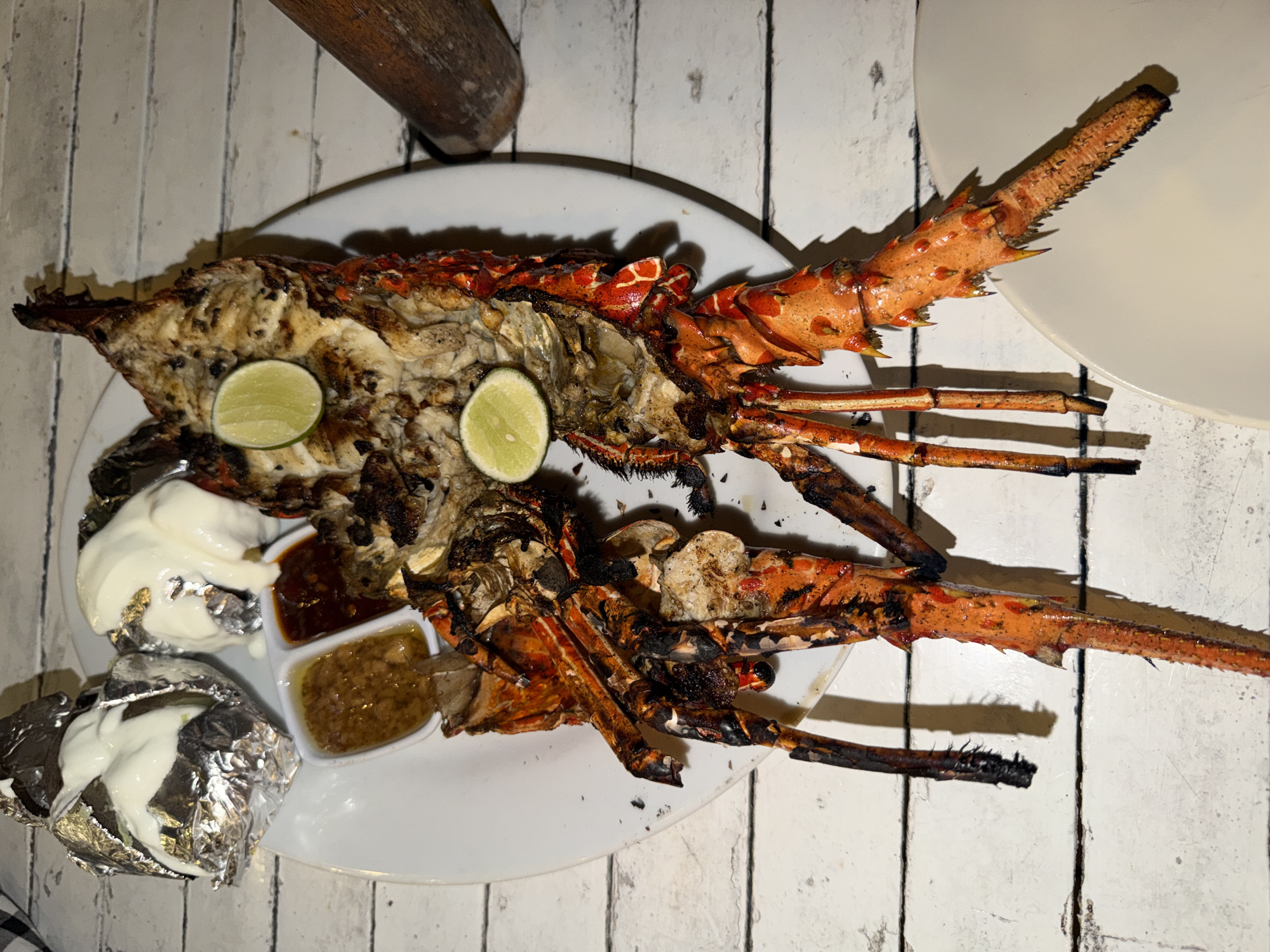A whole grilled lobster is presented on a white plate, seasoned with herbs and spices, and garnished with two lime wedges. The lobster is split lengthwise, revealing its succulent meat. Alongside the lobster, there are two small dipping sauces in separate compartments of a dish, one appearing to be a light-colored sauce and the other a reddish-brown sauce. To the upper left of the plate are two foil-wrapped items, each topped with a dollop of a creamy white sauce. The setting is an outdoor restaurant or beachside eatery, suggested by the weathered white wooden table and the presence of a rustic wooden tool, possibly a mortar and pestle, to the right of the plate. The overall lighting suggests it might be evening or a dimly lit environment. The location is Unknown, Indonesia.