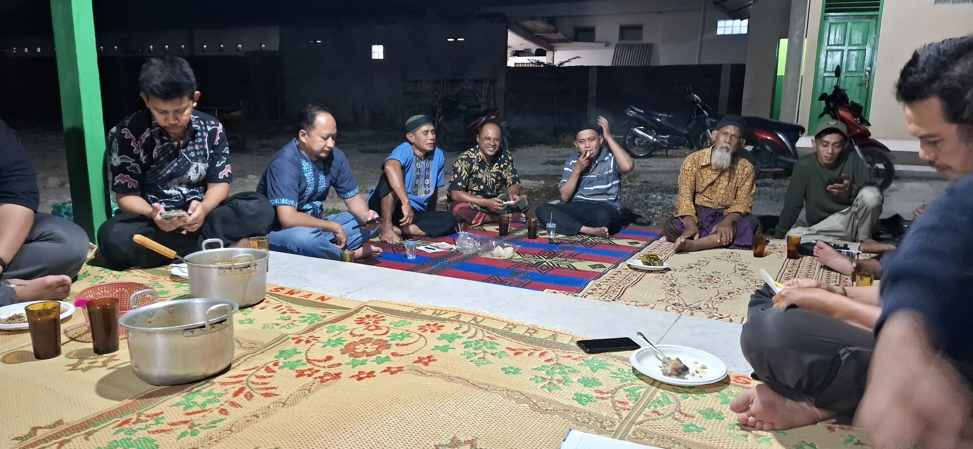 A group of seven men are gathered outdoors on woven mats in what appears to be a residential area in Wonorejo, Indonesia. The time of day is evening or night, indicated by the low light conditions and the presence of outdoor lighting fixtures in the background.
The men are seated on the ground, some of them cross-legged, in a semi-circle. They are dressed in casual attire, with some wearing traditional Indonesian clothing like sarongs. Several of the men are holding or interacting with their mobile phones. One man in the center is smoking.
In the foreground, there are two large metal cooking pots, several glasses, and plates with food. A red plastic basket is also visible. The mats are colorful and patterned. In the background, there are structures that resemble houses or buildings, as well as a motorcycle. The overall atmosphere appears to be relaxed and informal, possibly a gathering or meal among friends or family.