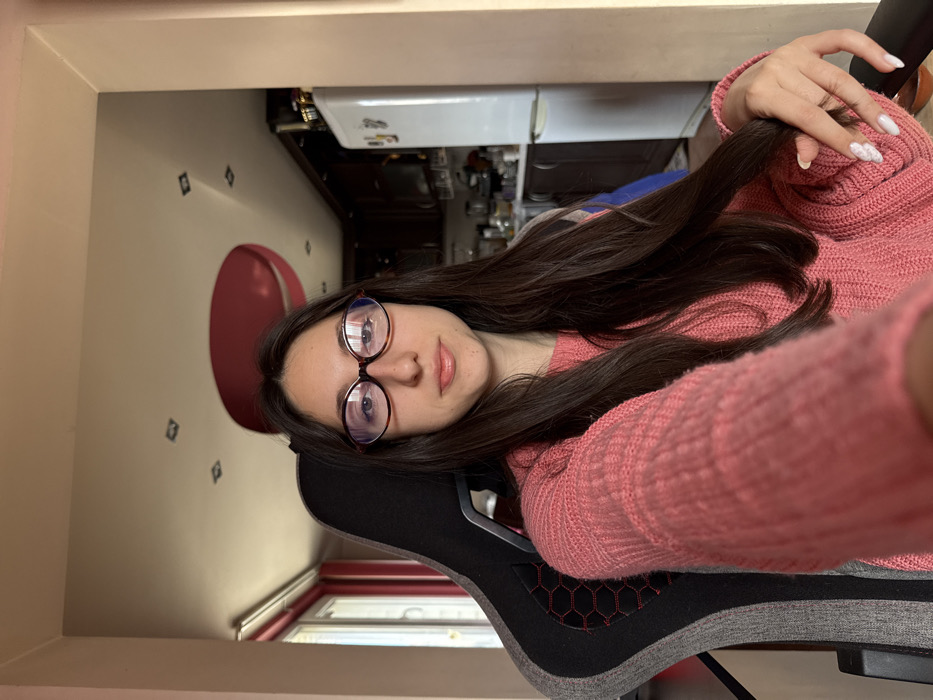 The image depicts an indoor residential scene in Galabovtsi, Bulgaria, focusing on a single female individual. She is seated centrally in a grey and black gaming chair with a hexagonal pattern on the backrest. The individual has long, dark brown hair, wears oval-shaped, dark-rimmed glasses, and a salmon-pink knitted sweater. Her expression is neutral as she looks directly at the camera. Her left hand, featuring white-marbled manicured nails, is visible holding a strand of her hair.
To her left, a window with a pink frame allows natural light into the room. To her right and in the background, a kitchen area is visible, featuring a white refrigerator, dark brown upper and lower cabinets, and a countertop with various items. The light-colored ceiling contains multiple square recessed lights and a prominent, large, circular, deep red decorative element. A doorway separates the seating area from the kitchen.