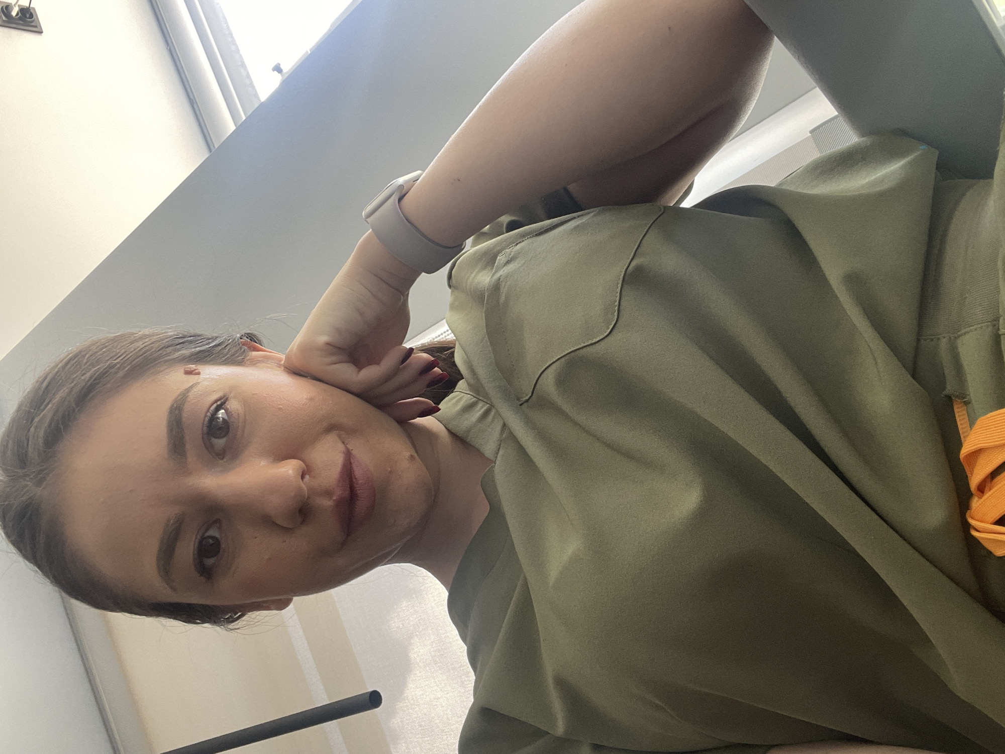 A selfie-style photograph shows a woman with light-brown hair and fair skin, wearing an olive-green garment. Her left hand is raised to her face, near her ear. She wears a light-grey smartwatch on her left wrist. Part of a window is visible in the upper-left corner of the frame, as well as a black bar in the lower-left corner. An orange cord or strap is visible on the right side of the frame. The location in the city of Sofia, Bulgaria is impossible to determine based on the visual information provided.