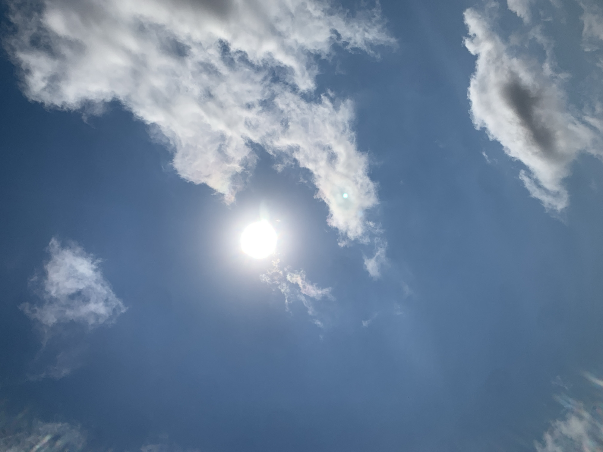 The image depicts a bright, sunny sky with scattered clouds over Khazar Raion, Azerbaijan. The sun is prominently featured, appearing as a bright, circular orb in the upper left quadrant of the image. Wispy white clouds are dispersed throughout the blue sky, with some appearing larger and more dense than others. The overall impression is of a clear day with some atmospheric haze.