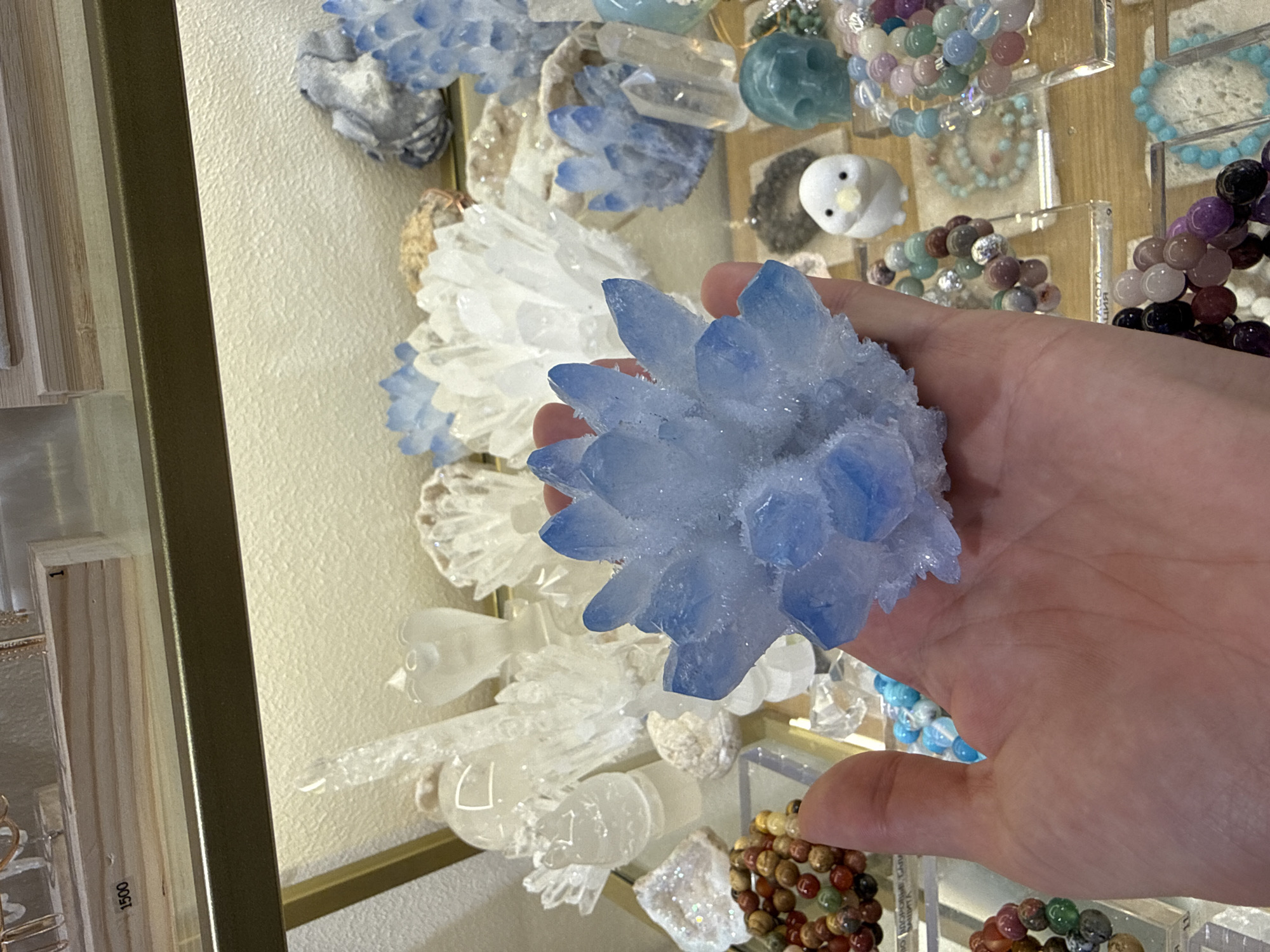 A hand holds a cluster of light blue crystals in the foreground of the image, in front of a display case filled with various gemstones and crystal formations. The display case contains a variety of items including bracelets made of colored beads, smaller crystal clusters, and carved stone objects. In the background, a price tag reading "1500" is visible on a wooden shelf. The overall setting appears to be a shop or market specializing in crystals and jewelry, likely in Saint Petersburg, Russia, given the Cyrillic text visible on some labels in the display case, such as "АРОМАТИЧЕСКИЯ" which translates to "AROMATIC."