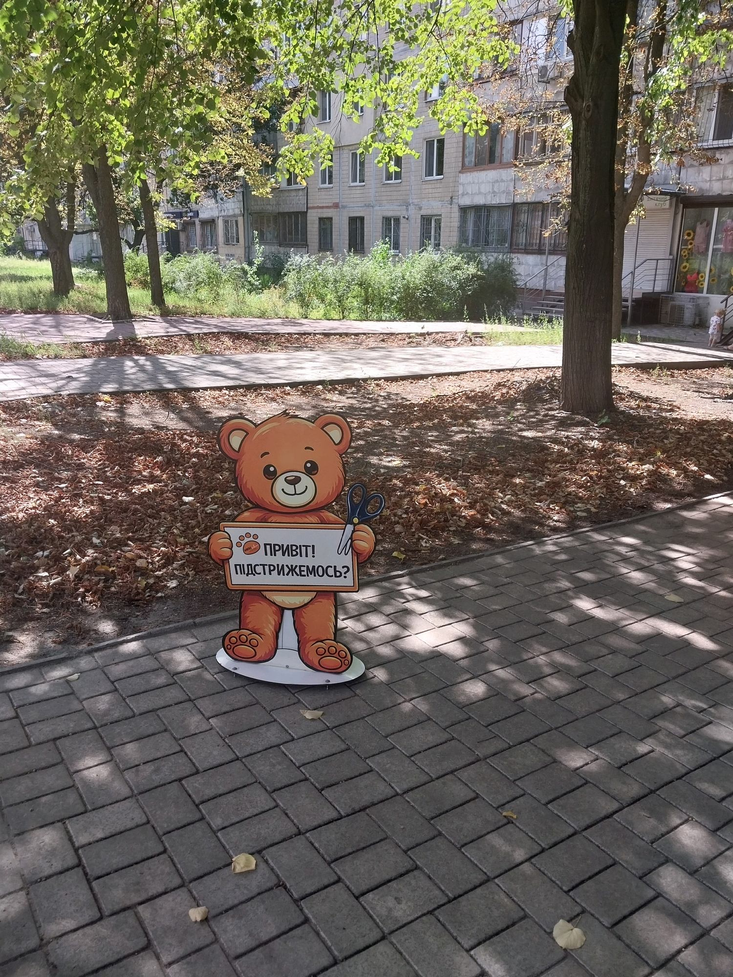 A cardboard cutout of a smiling teddy bear stands on a paved sidewalk in Dnipro, Ukraine. The bear, which is orange and brown, holds a white sign with text and paw prints. The text on the sign reads "ПРИВІТ! ПІДСТРИЖЕМОСЬ?" (Hello! Shall we get a haircut?). The bear is wearing blue scissors in its right paw. Behind the bear, the ground is covered in fallen leaves, and trees with green and yellow leaves provide shade. In the background, a multi-story residential building with multiple windows and balconies is visible. The sidewalk is made of grey rectangular pavers. The scene appears to be outdoors on a sunny day.