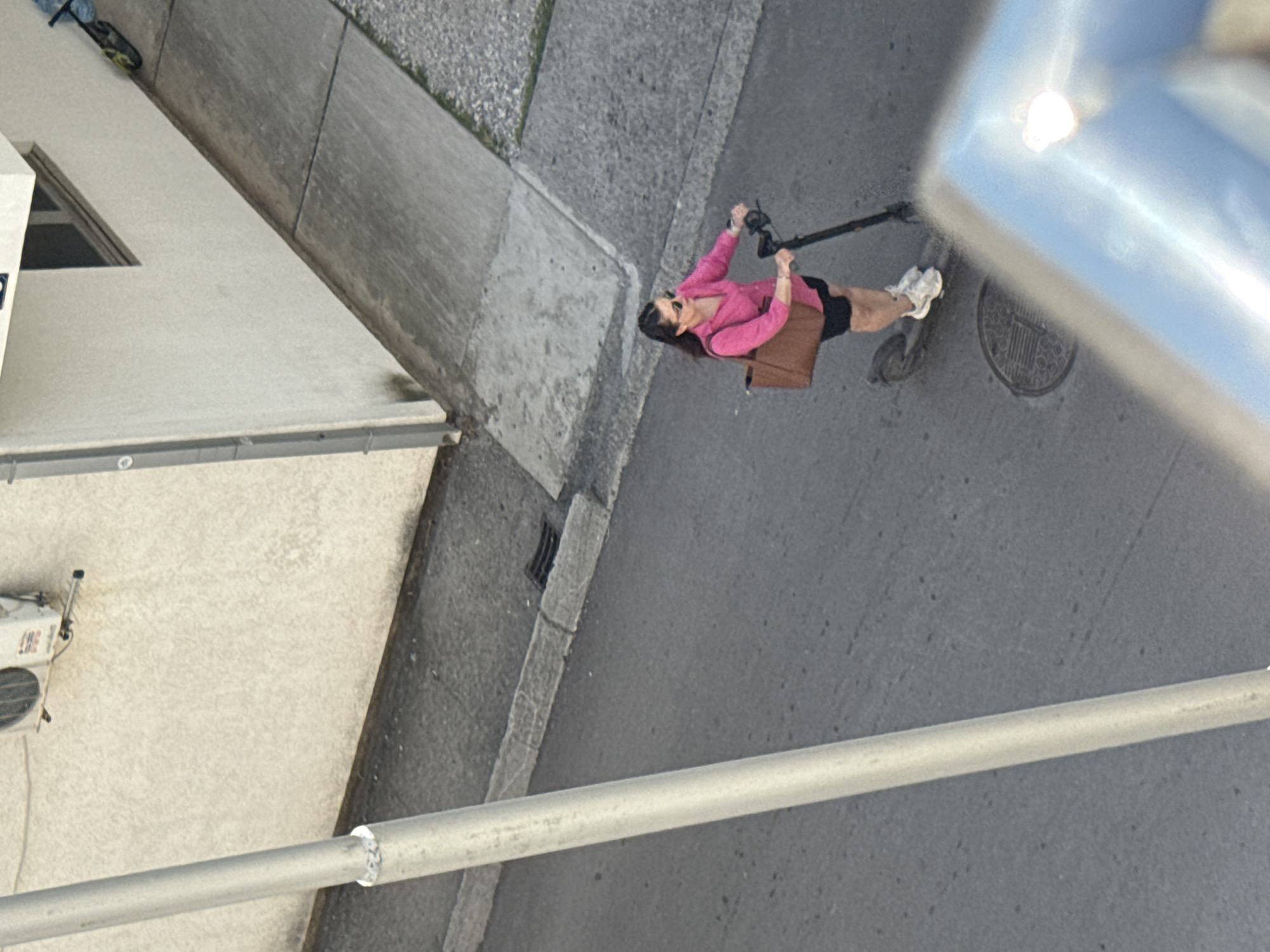 The image, taken from an elevated perspective, shows a woman riding an electric scooter on a city street. The woman is wearing a pink top, a black skirt or shorts, and white sneakers. She is also carrying a brown bag. A building is partially visible on the left side of the image. The street is made of asphalt, and there is a sidewalk visible along the building. A metal bar runs horizontally across the lower part of the image. A drain cover is visible on the street near the scooter. An air conditioning unit is visible on the side of the building. Given the architecture and climate, it's plausible this is the city of Budva, Montenegro.