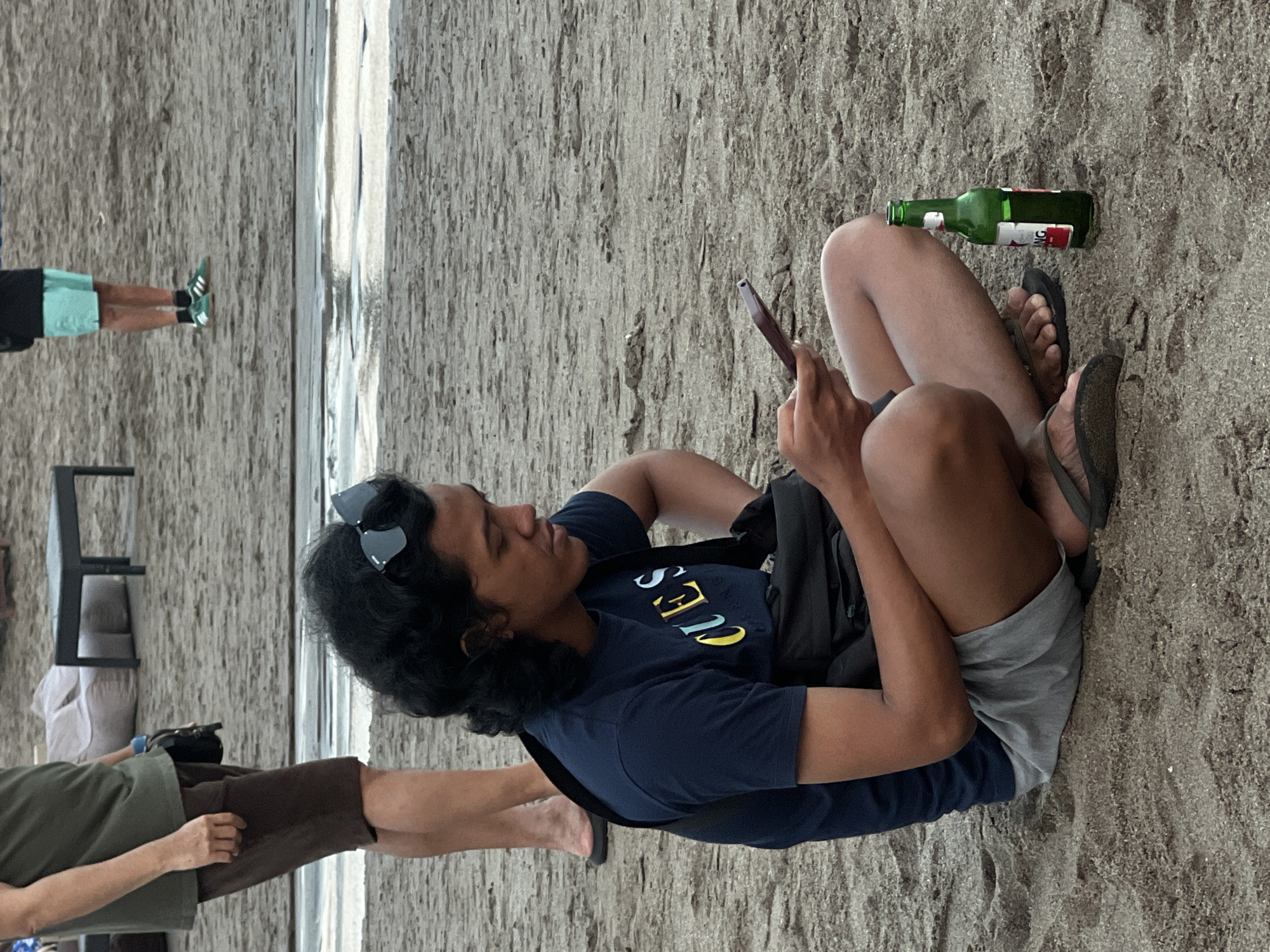 A young man with dark, curly hair is sitting on a sandy beach in North Kuta, Indonesia, looking at his phone. He is wearing a navy blue t-shirt with colorful lettering and gray shorts. A black bag is slung across his shoulder, and a green bottle of beer stands upright in the sand next to his right leg. Sunglasses are perched on top of his head. In the background, other beachgoers and furniture are visible. The sand is a light brown color with visible footprints and some wet patches.
