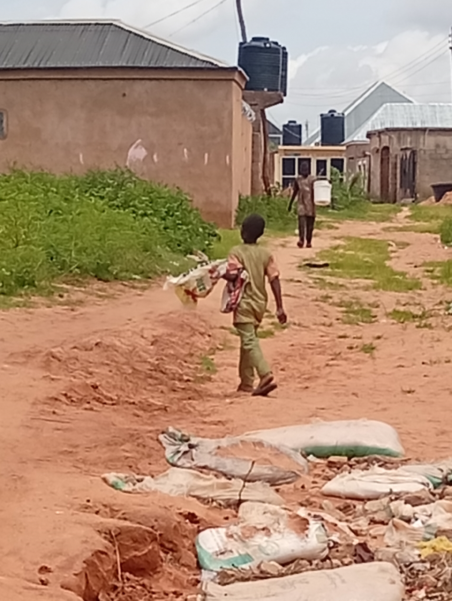 In Tumfure, Nigeria, a dirt path winds through a residential area with simple, unfinished buildings and scattered debris. Two young boys are seen walking along the path. The boy in the foreground, wearing a green shirt and pants, carries a white bag slung over his shoulder. Further down the path, a second boy, dressed in a dark shirt and lighter pants, walks carrying a white bucket. The environment appears dry and dusty, with sparse vegetation and piles of white sacks, possibly containing sand or cement, scattered around. Several large black water tanks are visible on rooftops in the background, suggesting water storage is common in the area. The sky is overcast with clouds.