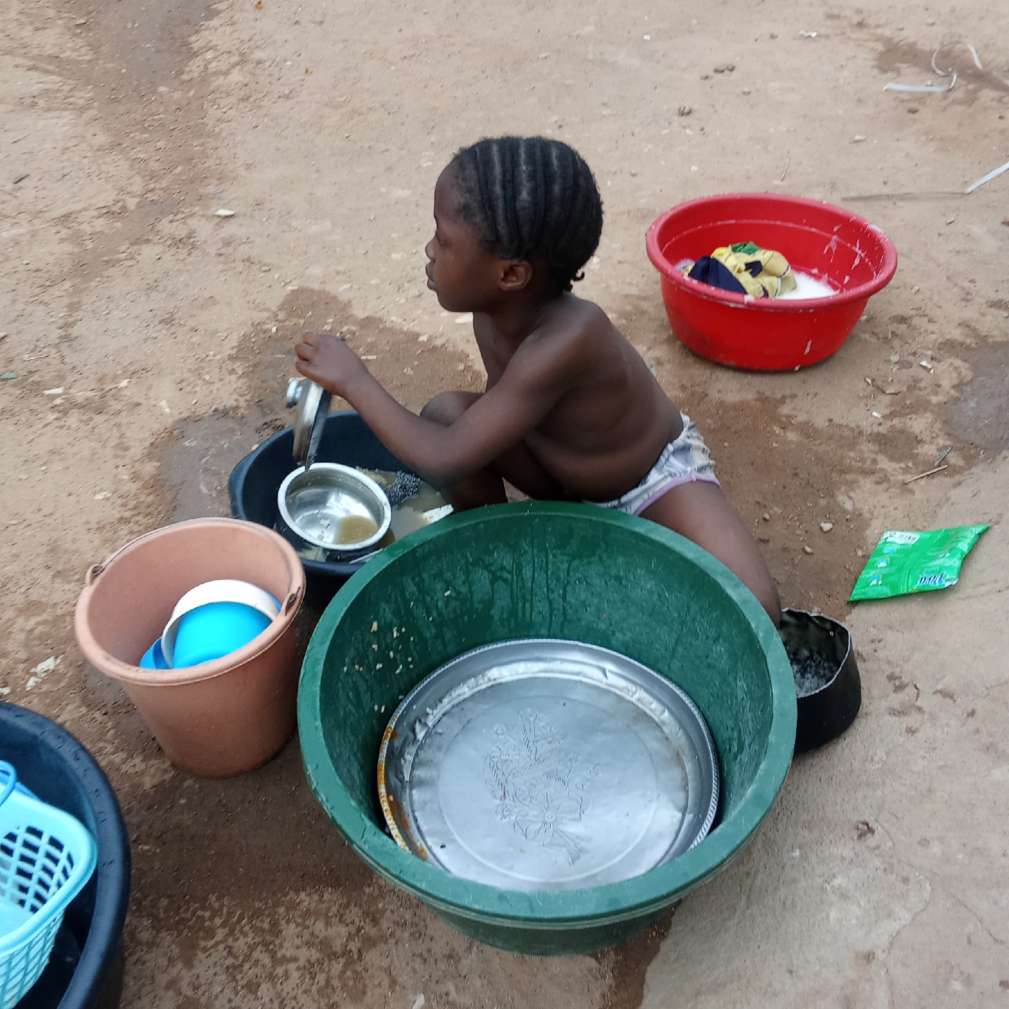 A young, dark-skinned child in Akwanga, Nigeria, washes dishes outdoors. The child, wearing patterned underwear, kneels among an array of buckets and basins containing soapy water and dishes. A red basin with soapy clothing sits nearby. A green packet lies on the ground to the right. The ground appears to be hard-packed dirt.