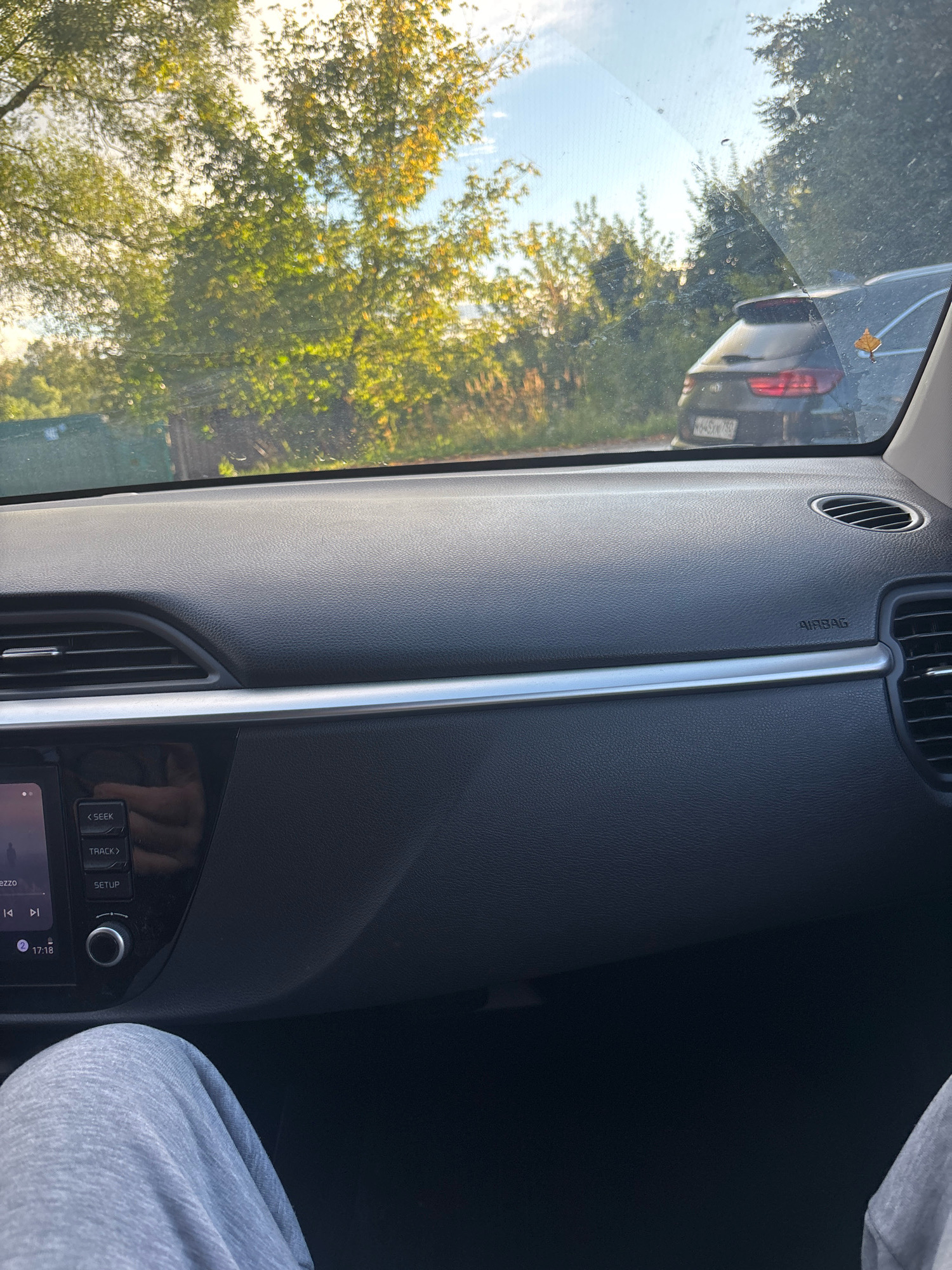 The image is taken from inside a car, looking out through the windshield. The view outside shows trees with green and yellowing leaves, suggesting it is either late summer or early autumn. A dark-colored SUV is visible on a road to the right, with a license plate that appears to be Russian. A single yellow leaf is stuck to the rear window of the SUV. The interior of the car is visible in the foreground, including the dashboard with air vents, a car stereo, and a person's legs wearing grey sweatpants. The overall scene suggests a journey through a suburban or rural area. There is no specific information about the exact location within the city of Sergiyev Posad, Russia, other than the presence of a Russian license plate.