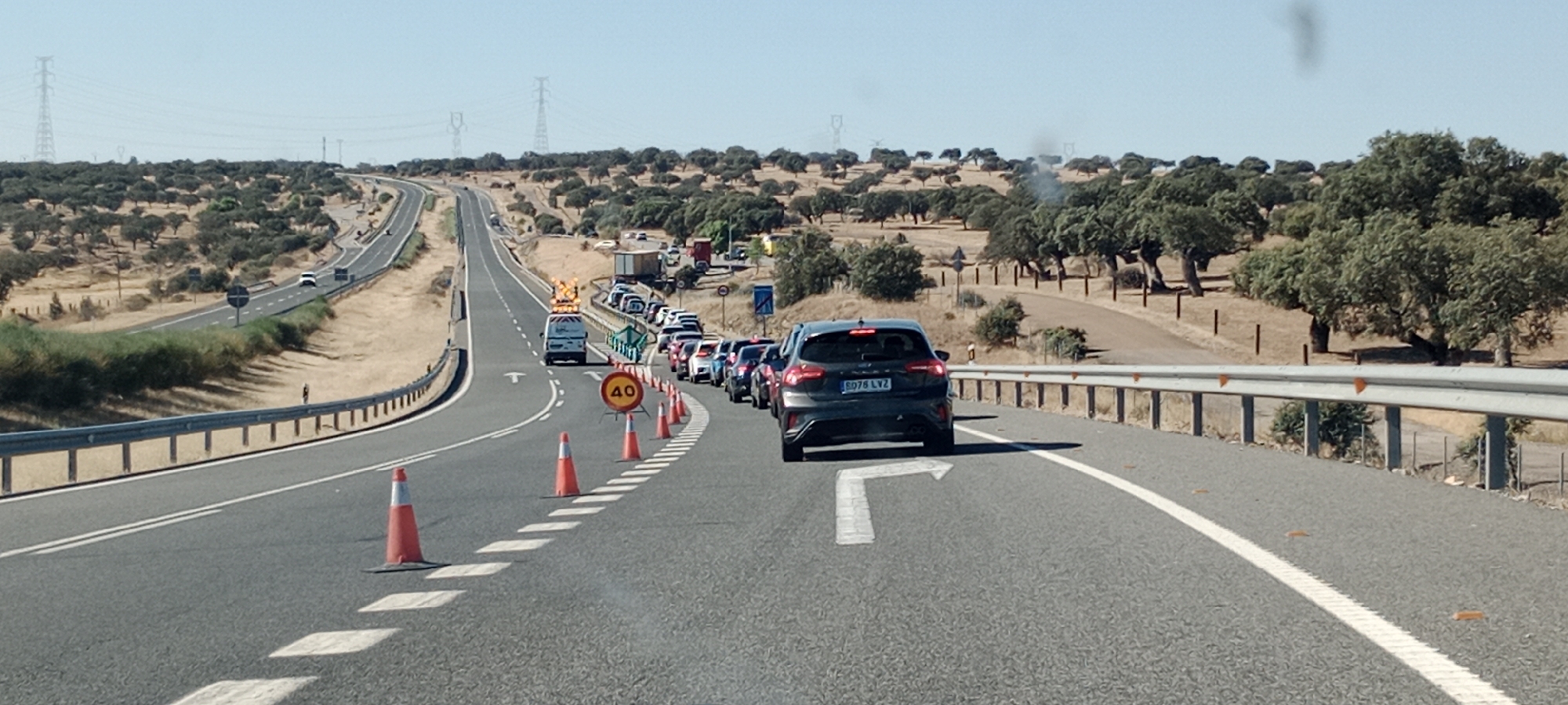 The image depicts a traffic jam on a highway in a rural, arid landscape with scattered trees. The day appears to be sunny and clear, suggesting warm weather. The road is paved, with multiple lanes separated by white lines. Traffic cones and a speed limit sign showing "40" are visible, indicating road work or some form of traffic control. A van with flashing lights and an orange warning sign on top is positioned at the front of the stalled traffic, likely related to the reason for the slowdown. Several cars are lined up behind it. In the distance, power lines stretch across the horizon, and the landscape is dotted with more trees. The overall scene suggests a frustrating delay for motorists. The license plate on the dark grey car in the foreground reads "B078 LVZ."