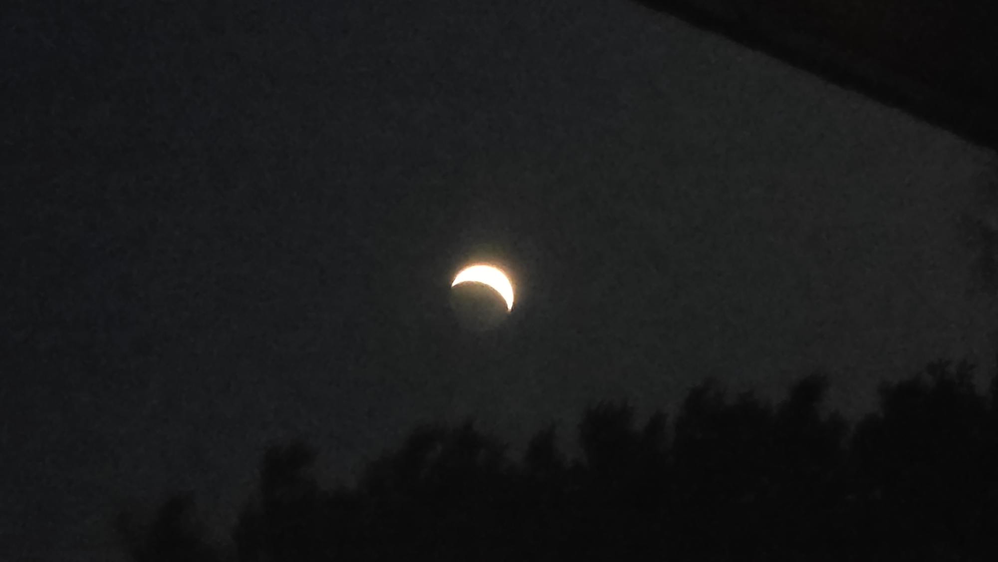 A crescent moon glows with a warm, orangey light in a dark, cloudy sky. A silhouette of a tree dominates the left side of the frame, its branches reaching upwards. A dark, angled shape, possibly a roofline or another structure, occupies the bottom right corner. The overall scene suggests a nighttime view, possibly from a residential area given the presence of trees and structures. The moon's illumination casts a faint glow on the surrounding clouds.