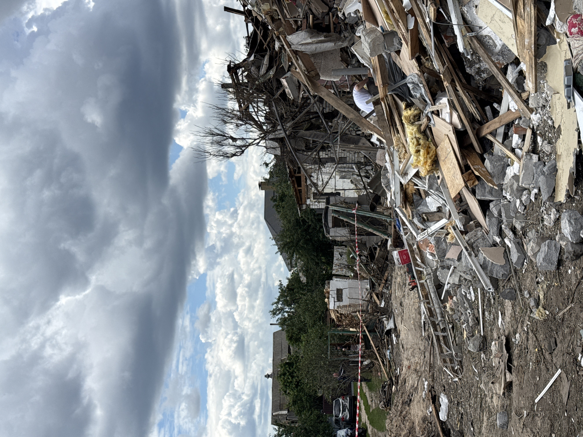 The image depicts a scene of significant destruction in a residential area of Kyiv, Ukraine. The foreground is filled with rubble from a collapsed structure, consisting of broken concrete, bricks, wooden planks, and insulation material. In the mid-ground, a person wearing a white shirt is visible amidst the debris, appearing to be searching or working within the wreckage. Further back, remnants of partially destroyed buildings are evident, including brick walls and twisted metal framing. Red and white tape is stretched across the scene, indicating a cordoned-off area. In the background, several residential houses with visible rooftops and trees are present, suggesting the destruction is within a populated neighborhood. The sky above is partly cloudy with a mix of dark storm clouds and patches of blue sky. Cars are parked on a dirt path to the left of the frame, and a large pile of debris obscures much of the immediate surroundings.