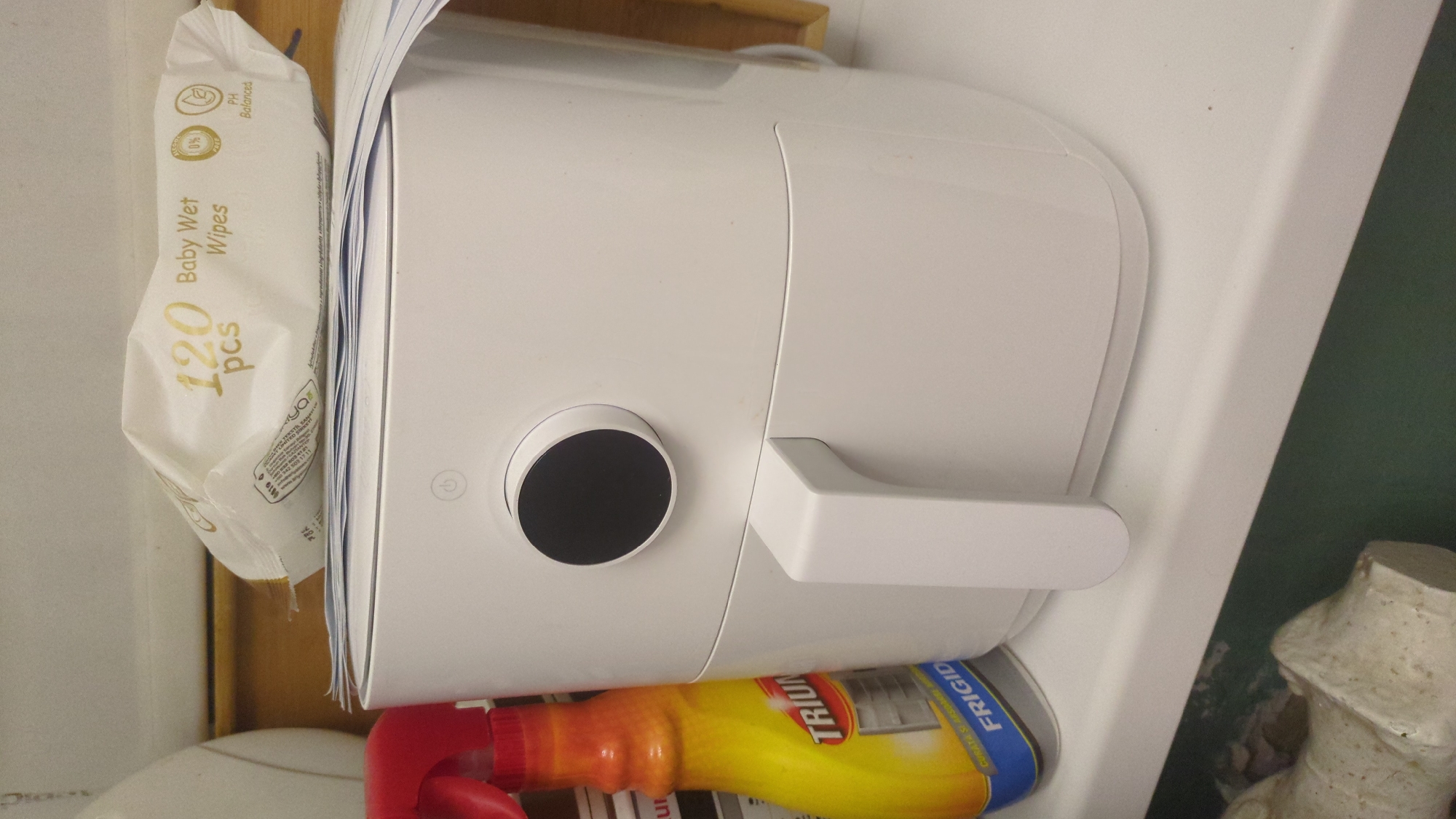 A white air fryer is positioned on a white shelf. To the left of the air fryer is a yellow bottle with a red spray nozzle, likely a cleaning product. On top of the air fryer, partially obscuring it, is a package of "Baby Wet Wipes" that contains "120 pcs." The background appears to be an indoor setting with shelves or cabinets. The image is a still photograph and does not depict any specific location in Chișinău, Moldova, nor does it show any people or actions. The focus is on the objects arranged on the shelf.