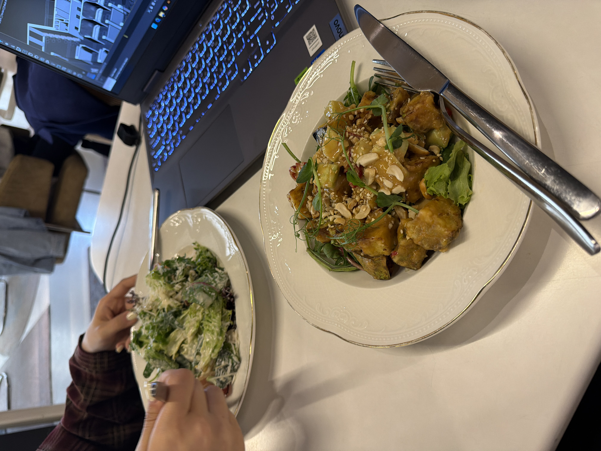 The image presents a detailed tabletop view within an indoor establishment, likely a cafe or restaurant. In the foreground-left, a person's hands, wearing a plaid shirt, are seen holding a fork to eat from a white plate containing a Caesar-style salad topped with dressing and grated cheese. The person's left hand has white-painted fingernails. To the right, a dark grey Lenovo laptop with a blue-backlit keyboard is open, displaying a screen with a 3D architectural rendering or CAD software interface. In the immediate foreground-right, another white plate with a gold rim and an embossed pattern holds a main course dish consisting of diced, glazed pieces of food (possibly vegetables or protein), garnished with chopped peanuts, sesame seeds, and microgreens. A silver knife and fork rest on the table beside this plate. The table surface is white. The background is blurred, revealing parts of another individual dressed in a dark blue top and brown seating. No identifiable specific location within Yerevan, Armenia, is discernable from the visual information provided.