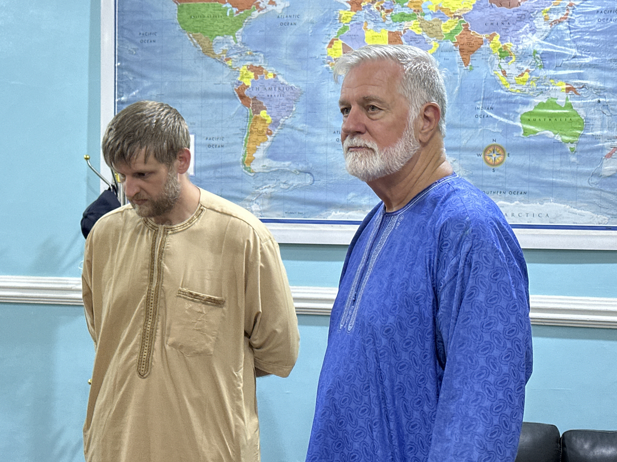 Two men are standing in front of a world map. The man on the left is wearing a tan robe with gold trim. He has short brown hair and a beard. The man on the right is wearing a blue robe with a patterned design. He has gray hair and a white beard and is looking to the right. The wall behind them is painted light blue, and there is a white molding at the top of the wall. The world map on the wall shows continents and oceans. There are no visible signs that this scene is located in Television, Nigeria.