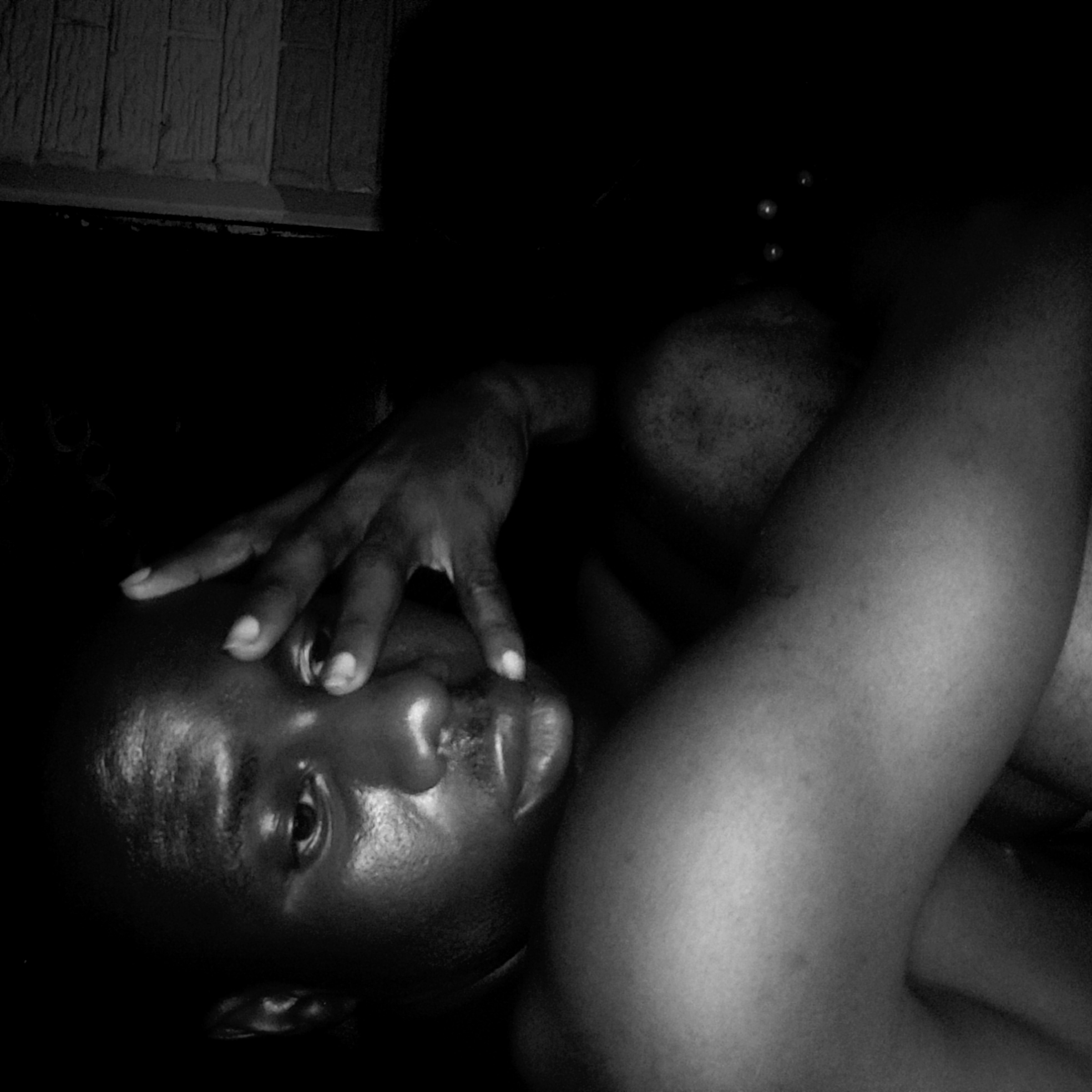 The black and white image depicts a shirtless Black male lying on his side. His head is slightly tilted, and his right hand is raised, with his fingers partially covering his face. His left arm is crossed over his body. The environment appears dark, with some indistinct textures in the background, suggestive of a wall. No specific details confirm the location as Narayi, Nigeria.