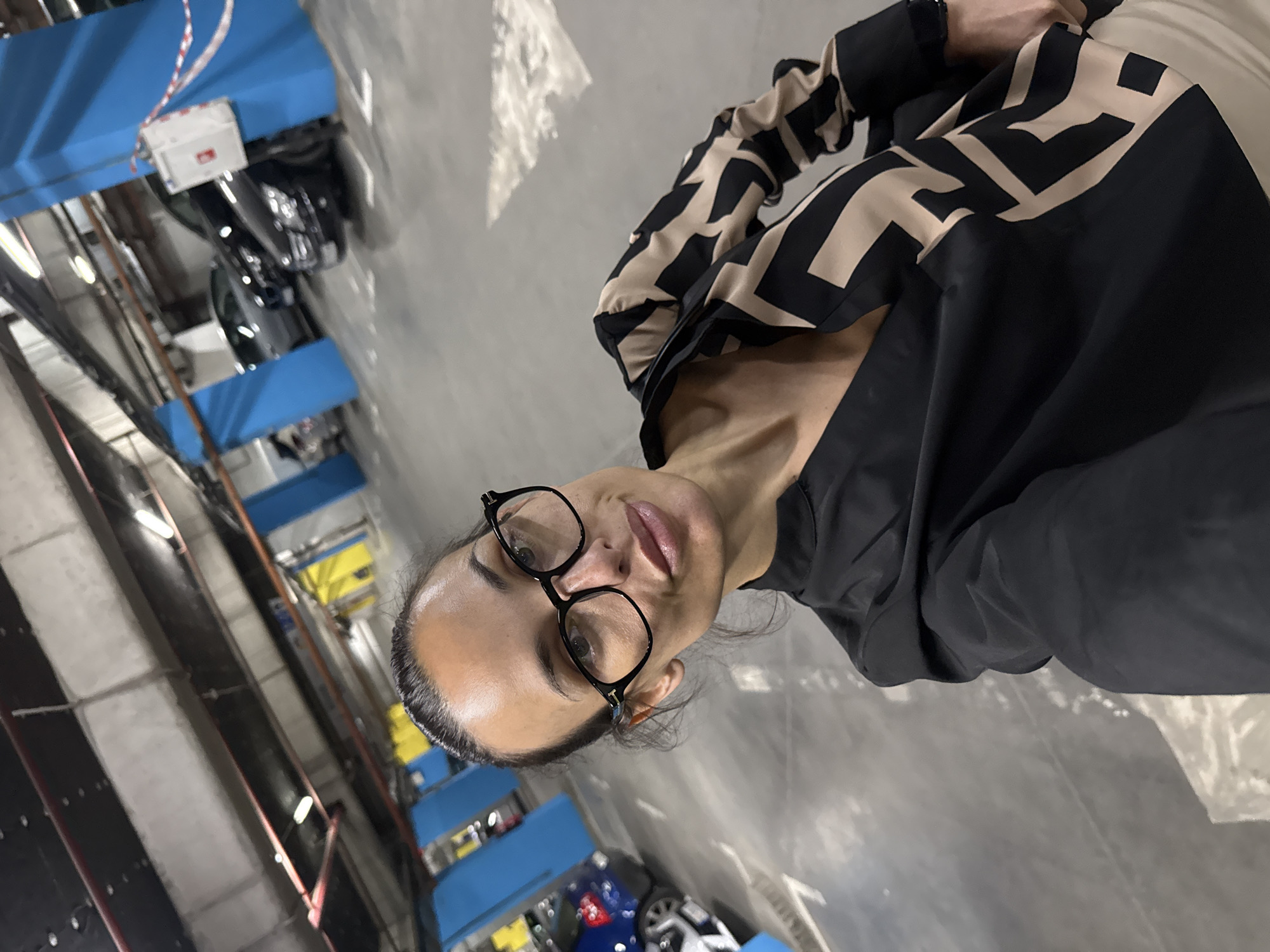 The image captures a woman posing for a photo in what appears to be an indoor parking garage or vehicle storage facility, likely located in Pleven, Bulgaria. The primary subject, a woman with dark hair pulled back and wearing black-framed eyeglasses, is looking directly at the camera with a neutral to slightly smiling expression. She is wearing a black top or jacket featuring a distinctive beige/tan geometric pattern on the sleeves and upper body.
The background shows an industrial-style environment characterized by concrete walls and pillars. Prominent blue metal structures, possibly components of a multi-level parking system or vehicle lifts, are visible on the left side of the frame. Several vehicles, including parts of dark-colored cars and a motorcycle, are discernible in the background, positioned on or near these structures. The area is illuminated by artificial fluorescent lighting. A small white box with some red markings and partially visible, indistinct text is attached to one of the blue metal structures. The setting suggests a functional space dedicated to vehicle storage or maintenance.