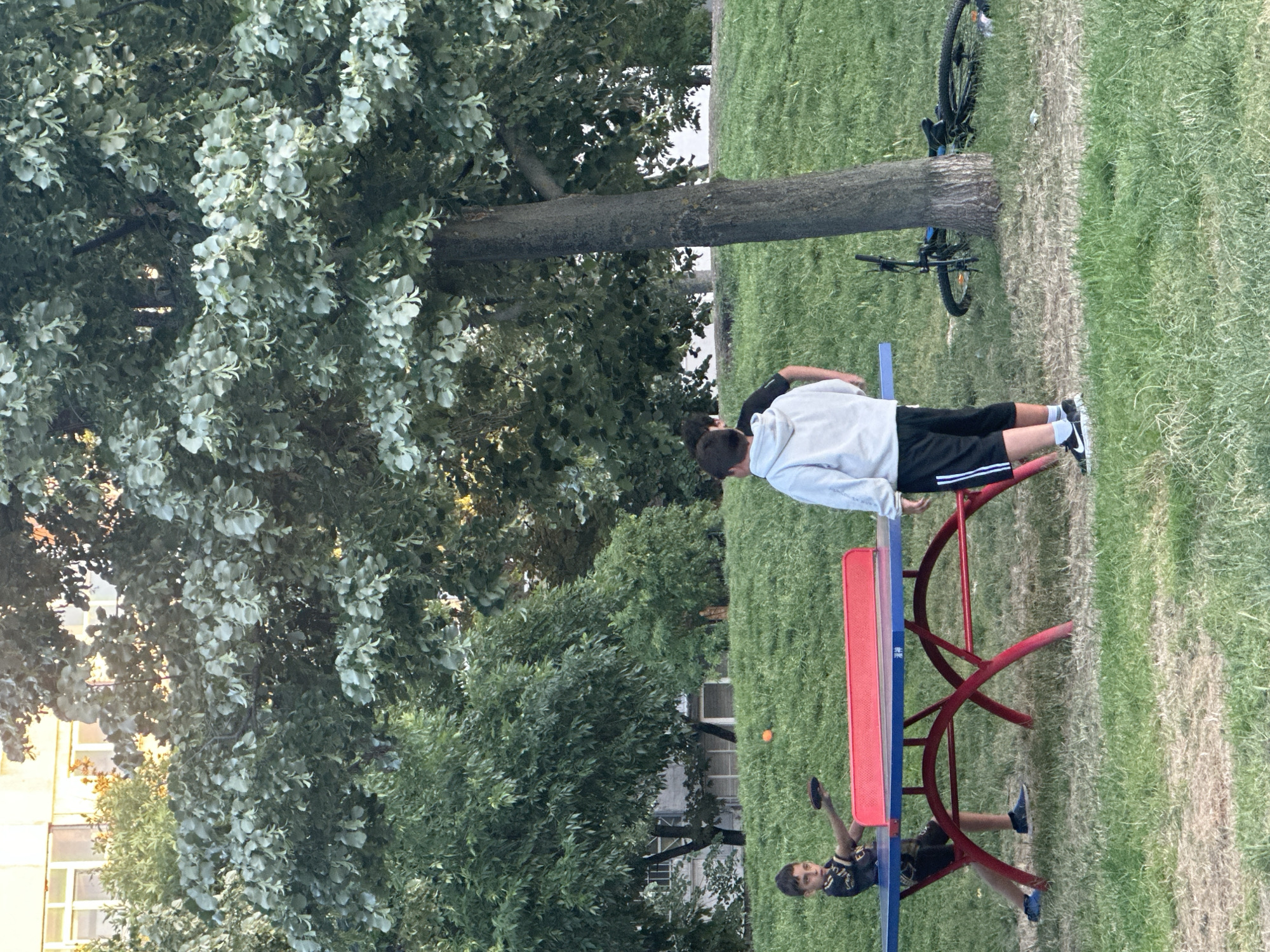 In a park in Sofia, Bulgaria, two young boys are playing table tennis. A third boy stands nearby. The table is blue with a red net and red legs. A bicycle lies on the grass to the right of the table. Lush green trees and grass fill the background. A building is visible behind the trees. The lighting suggests it is daytime.