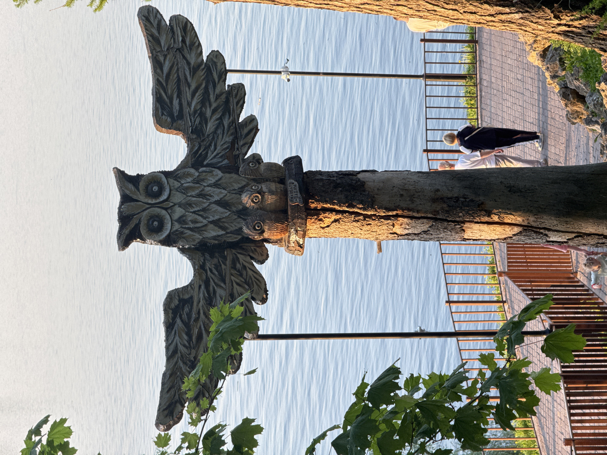 A wooden owl sculpture with spread wings, adorned with smaller owl carvings on the trunk, stands on a bank of the Kama River in the city of Krasny Klyuch, Russia. The sculpture is positioned near a sidewalk with a metal railing where two people are visible: one is wearing a white shirt and white pants, the other is wearing a black jacket. A security camera is installed above the sculpture. There are trees visible on either side of the image.
