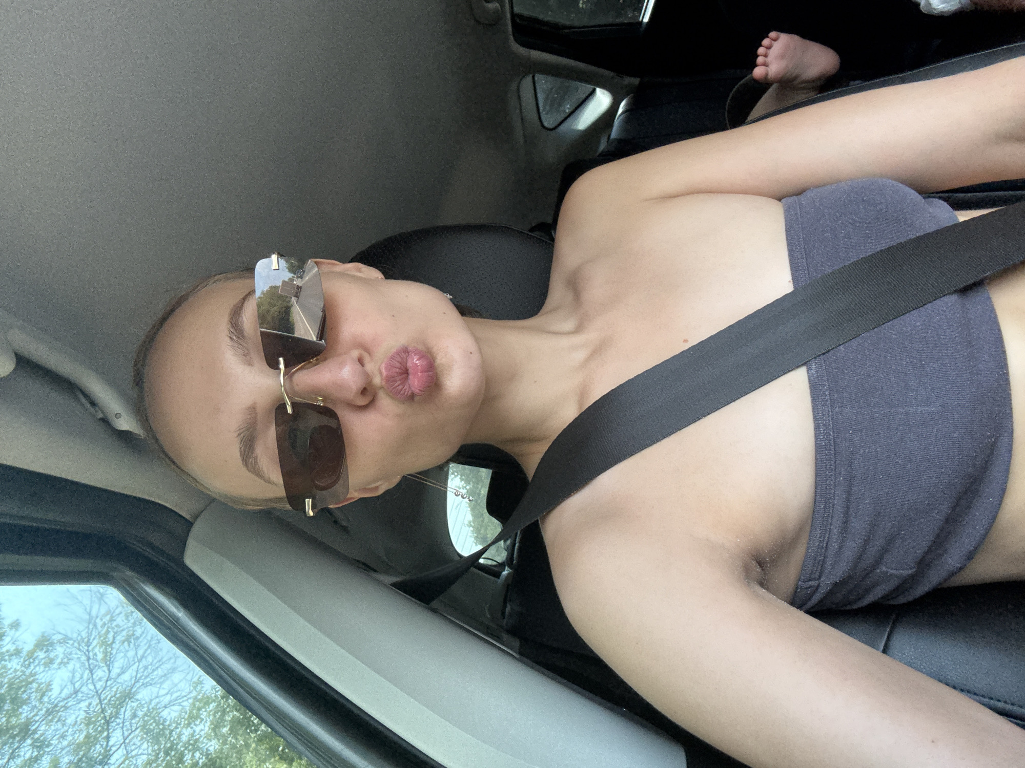 A young woman with blonde hair pulled back is in the driver's seat of a car, wearing sunglasses and a grey strapless top. She is puckering her lips for a selfie. A seatbelt is visible across her chest. In the background, a child's feet are visible in a car seat. The image appears to be taken during the daytime, with trees and the sky visible through the car window, suggesting an outdoor, possibly rural, location. The specific location within Izhevsk, Russia, cannot be determined from the image alone.