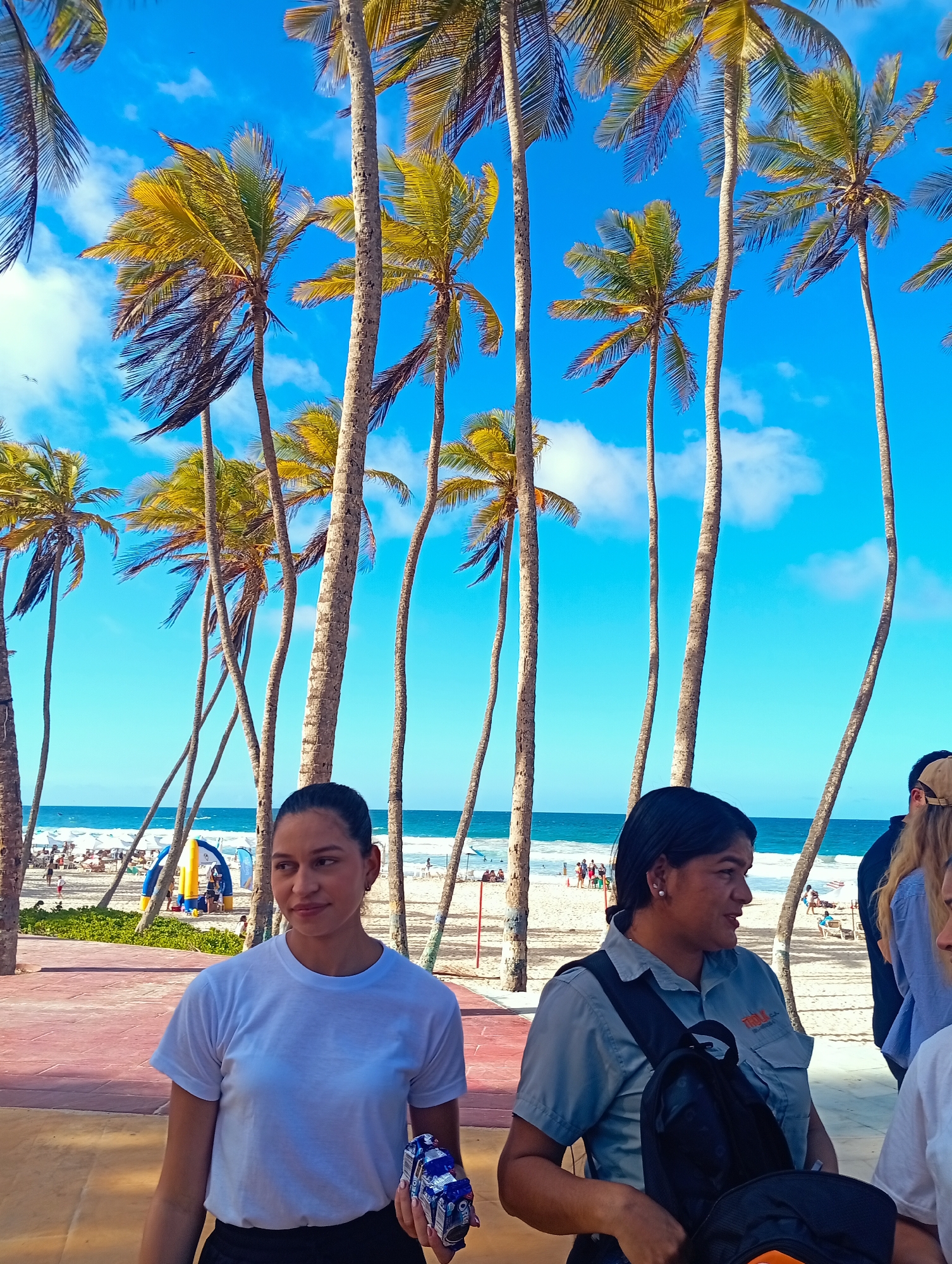 The image depicts a scene at Playa El Agua, Venezuela, with tall palm trees dominating the backdrop. In the foreground, two women are visible: one in a white t-shirt holding a package, and another in a blue uniform shirt with a black backpack. Behind them, a sandy beach stretches towards the ocean, dotted with beachgoers, umbrellas, and a partially visible blue inflatable structure. Other people are near the woman in the blue uniform. The sky is bright blue with scattered white clouds.