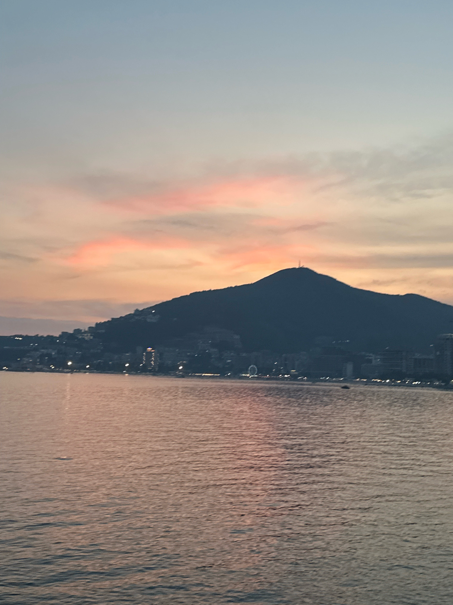 A silhouette of a mountain with buildings and lights at its base sits behind a body of water. The water reflects the muted colors of the sky, which transitions from a pale blue at the top to soft pink and orange hues lower down, indicating either sunrise or sunset. A thin antenna is visible at the very peak of the mountain. The shoreline is lined with structures, some of which are illuminated, suggesting a coastal town or city. The water is calm, with gentle ripples on its surface. This scene is consistent with the view of the Adriatic Sea with the mountain of `Spas` in the background, a prominent landmark overlooking the city of Budva, Montenegro.