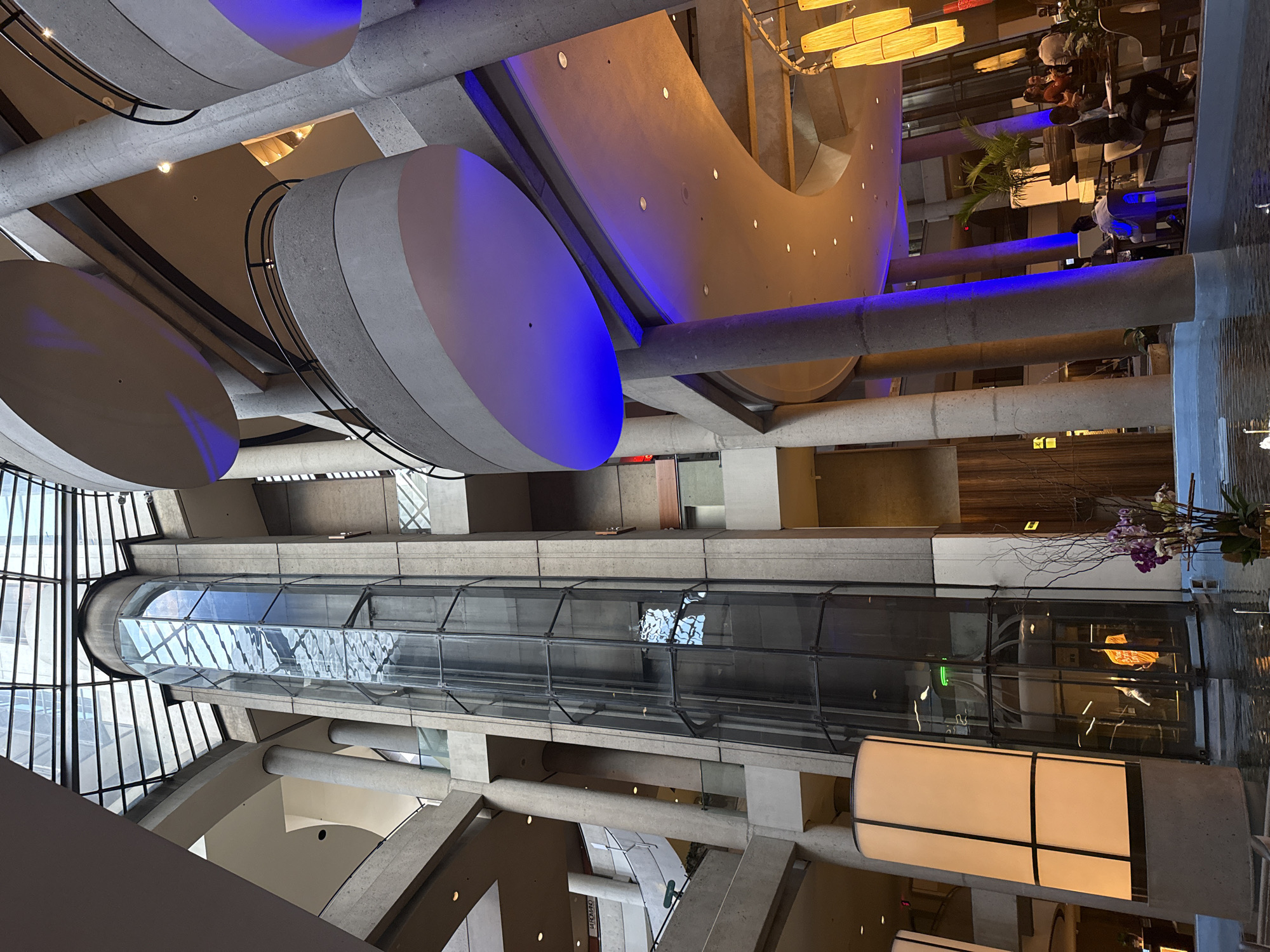 The image shows the interior of a multi-level atrium with a central glass elevator. The architecture features concrete columns and circular balconies, illuminated by blue and orange lights. In the foreground, a water feature with a plant and flowers is visible. Towards the right, a restaurant or cafe area is set up with tables and chairs, where several people are seated, including a man in a suit at a table. The upper levels of the atrium have a glass ceiling that allows natural light to enter, and there are visible architectural elements of the building's structure. The scene suggests an indoor public space, possibly a hotel lobby or a commercial building with amenities. The location is identified as the Dorothy Chandler Pavilion in Los Angeles, California, United States of America.