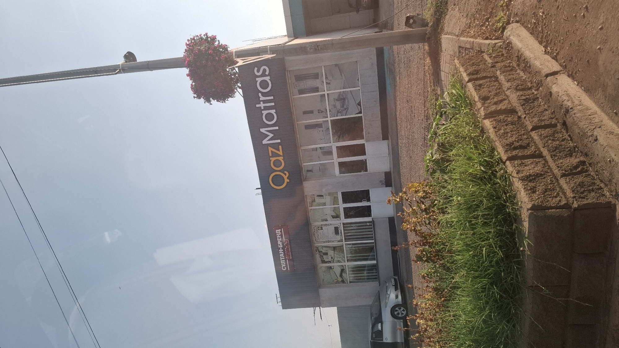 The image depicts the exterior of a commercial building identified as "QazMatras" located in Shymkent, Kazakhstan. The building has a modern facade with large windows displaying interior scenes, likely of mattresses or bedroom furniture. The "QazMatras" logo is prominently displayed on the building's dark upper section. To the left of the "QazMatras" signage, there is another sign that reads "СУЛТАН-БРЕНД". A silver sedan is parked in front of the building. In the foreground, there is a raised flower bed with lush green grass and some dry plants, constructed with interlocking concrete blocks. A tall pole with a hanging flower basket is visible to the right of the building. The sky is clear and blue, suggesting daytime. The scene is likely captured from a moving vehicle, as indicated by the slight blur and the angle.