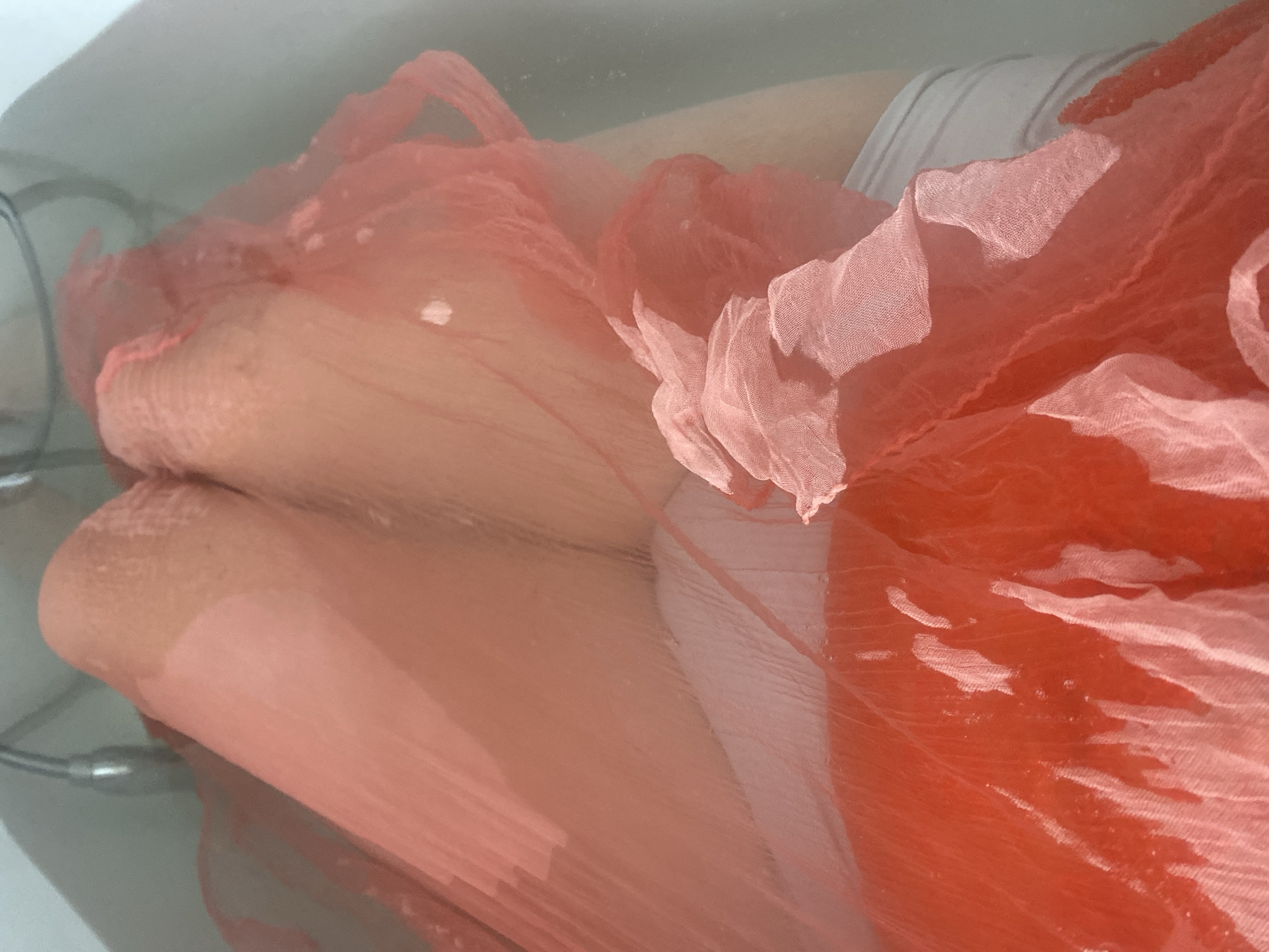 The image depicts a person's legs submerged in a bathtub. The legs are covered in a sheer, coral-colored fabric, which appears to be wet and draped over them. Underneath the coral fabric, white underwear is visible. The bathtub itself is white and appears to be a standard bathtub, with a chrome fixture visible at the top of the frame, suggesting a showerhead or faucet. The water in the tub is clear, with some small bubbles and ripples visible on the surface. The lighting is soft and diffused, creating a slightly hazy or ethereal effect. The scene suggests a moment of relaxation or personal care. The location in Novi Petrivtsi, Ukraine, is not visually identifiable from the image itself, as the scene is entirely contained within the bathtub.