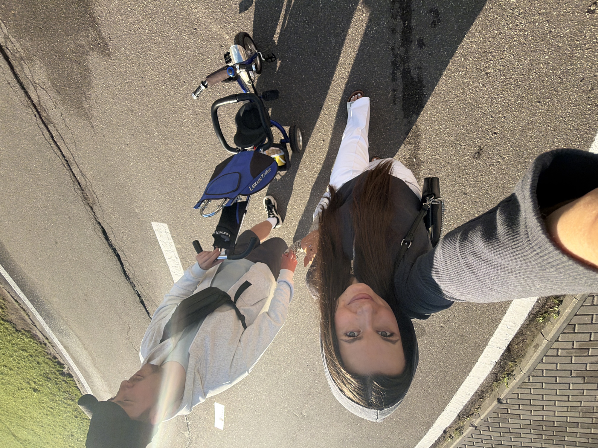 In Odintsovo, Russia, a young woman with long brown hair and a grey hooded jacket is taking a selfie outdoors on an asphalt road. She is smiling at the camera, and her arm is extended. Behind her, a young man wearing a black baseball cap and a light grey hooded jacket is looking away from the camera, standing next to a blue and black tricycle. The tricycle has a blue bag on the back with the words "Lexus Trike" printed on it. The background consists of a grassy area and a paved sidewalk. The lighting suggests it is daytime, likely late afternoon due to the long shadows.
