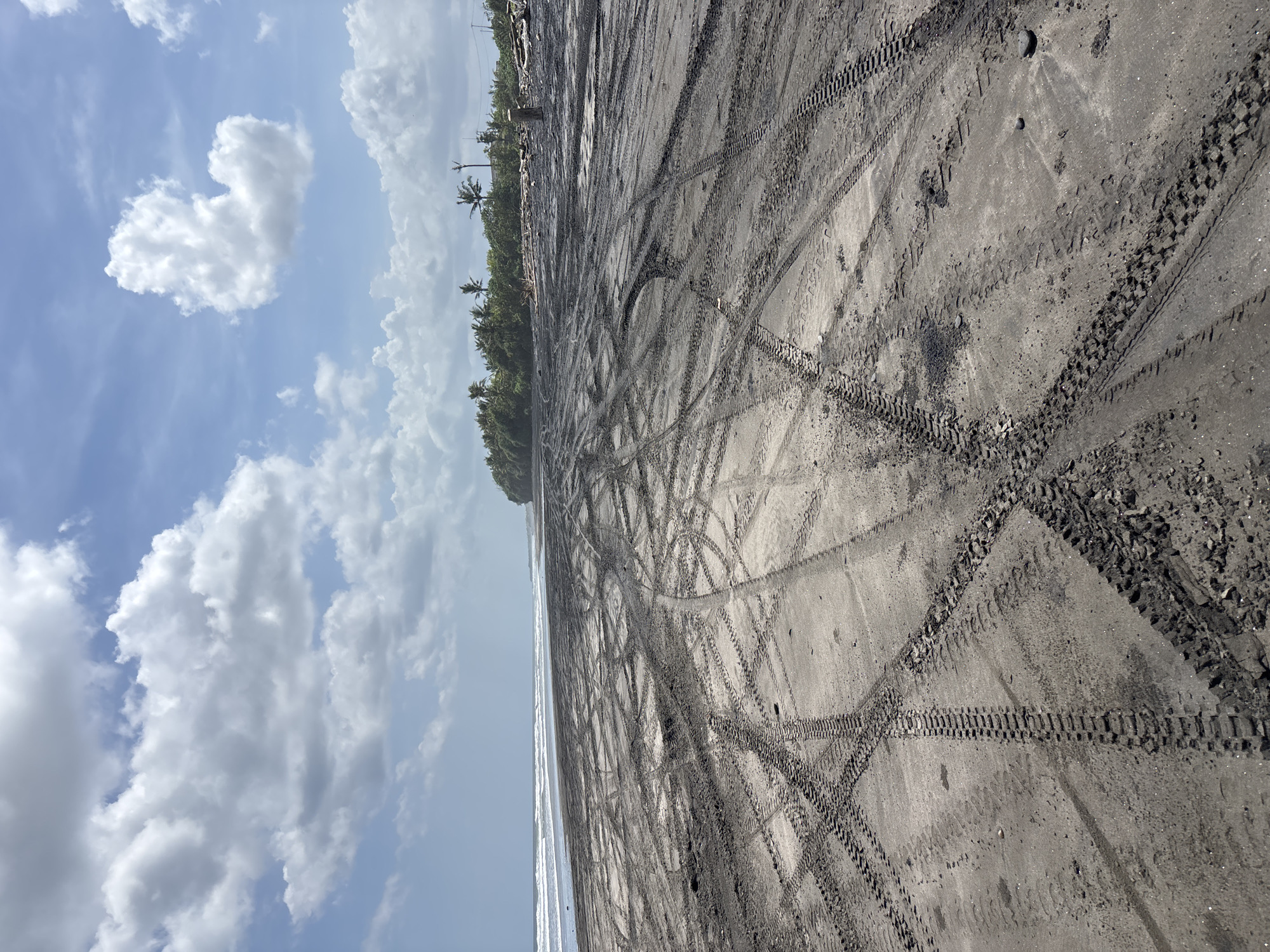 The image shows a beach in Kerambitan, Indonesia, with dark sand covered in tire tracks. A line of trees is present along the edge of the beach, separating it from inland areas. The ocean is visible along the left side of the image, and the sky is cloudy. No people or other subjects are visible in the scene.