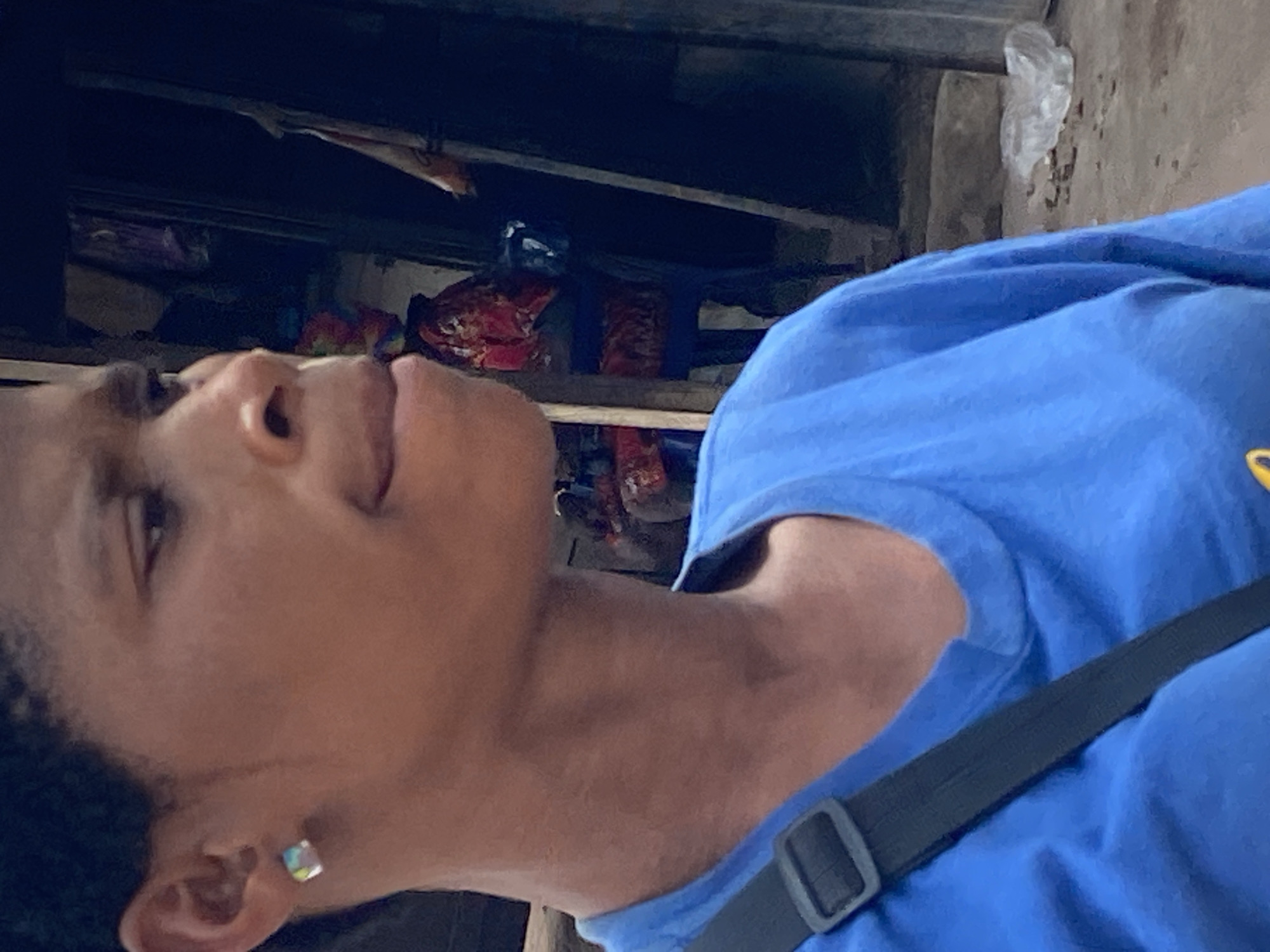 A young Black woman is shown in a medium close-up, from the shoulders up, in what appears to be a city setting. She is wearing a bright blue t-shirt and a black strap from a bag is visible across her chest. She has a small, square, iridescent earring in her left ear. Her head is turned to the right, and she is looking slightly downward and to the side, with her mouth partially open. The background is blurred and dark, with hints of fabric and structures suggesting an outdoor market or stall area. The overall lighting is dim, casting shadows and highlighting the contours of her face and neck. This scene is located in Nguzu Edda, Nigeria.