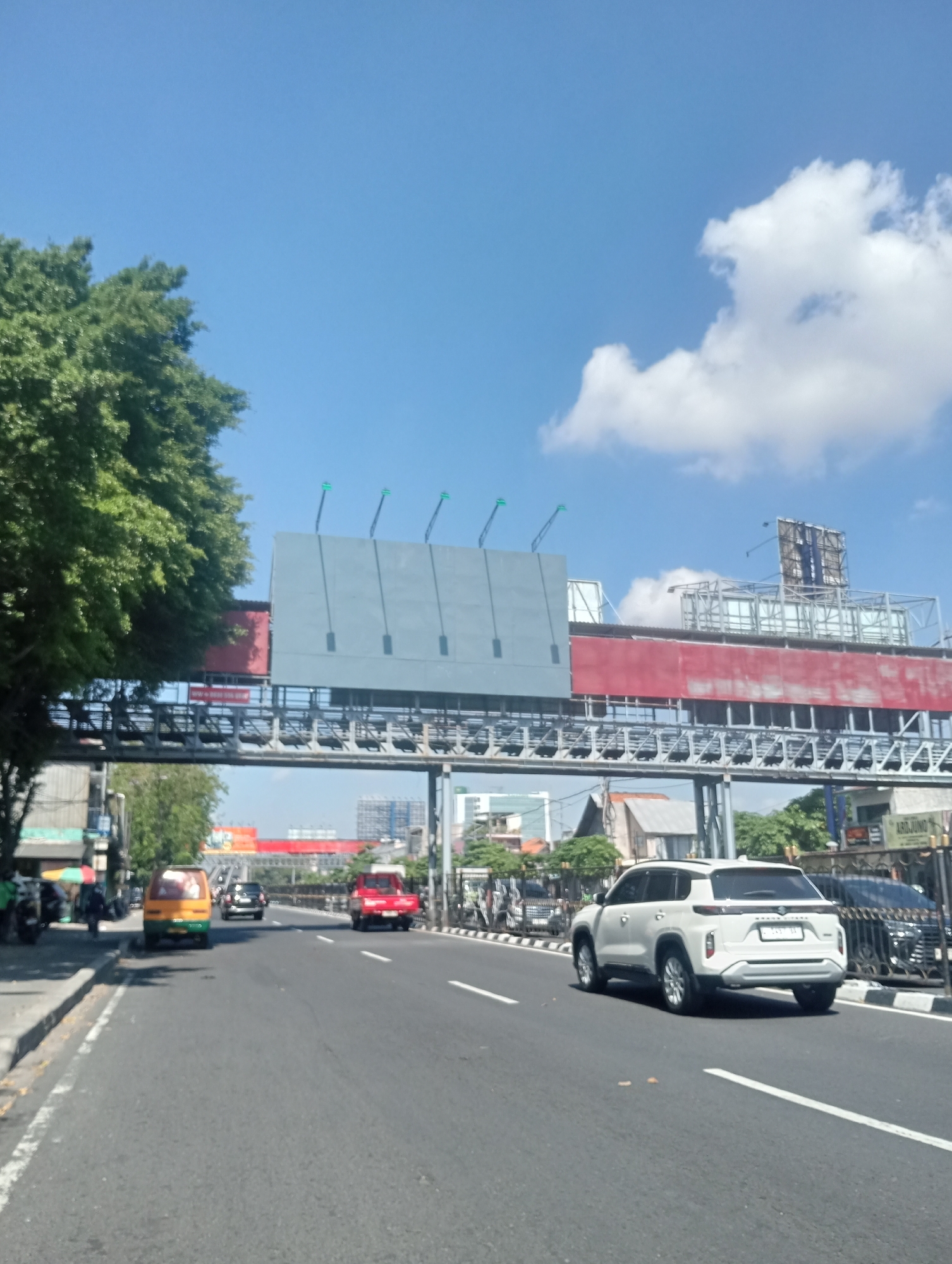 A wide, sun-drenched asphalt road stretches into the distance under a clear blue sky dotted with white clouds. On the left side of the road, a lush green tree provides shade, bordering a sidewalk with a few parked motorcycles and a small green and yellow vehicle. Further down the road, traffic flows, with a red car and a white SUV prominently visible. Overhead, a large, grey billboard structure spans across the road, supported by a metal framework. The surrounding cityscape features a mix of buildings and trees, suggesting an urban environment. The overall scene conveys a typical day in Surabaya, Indonesia.