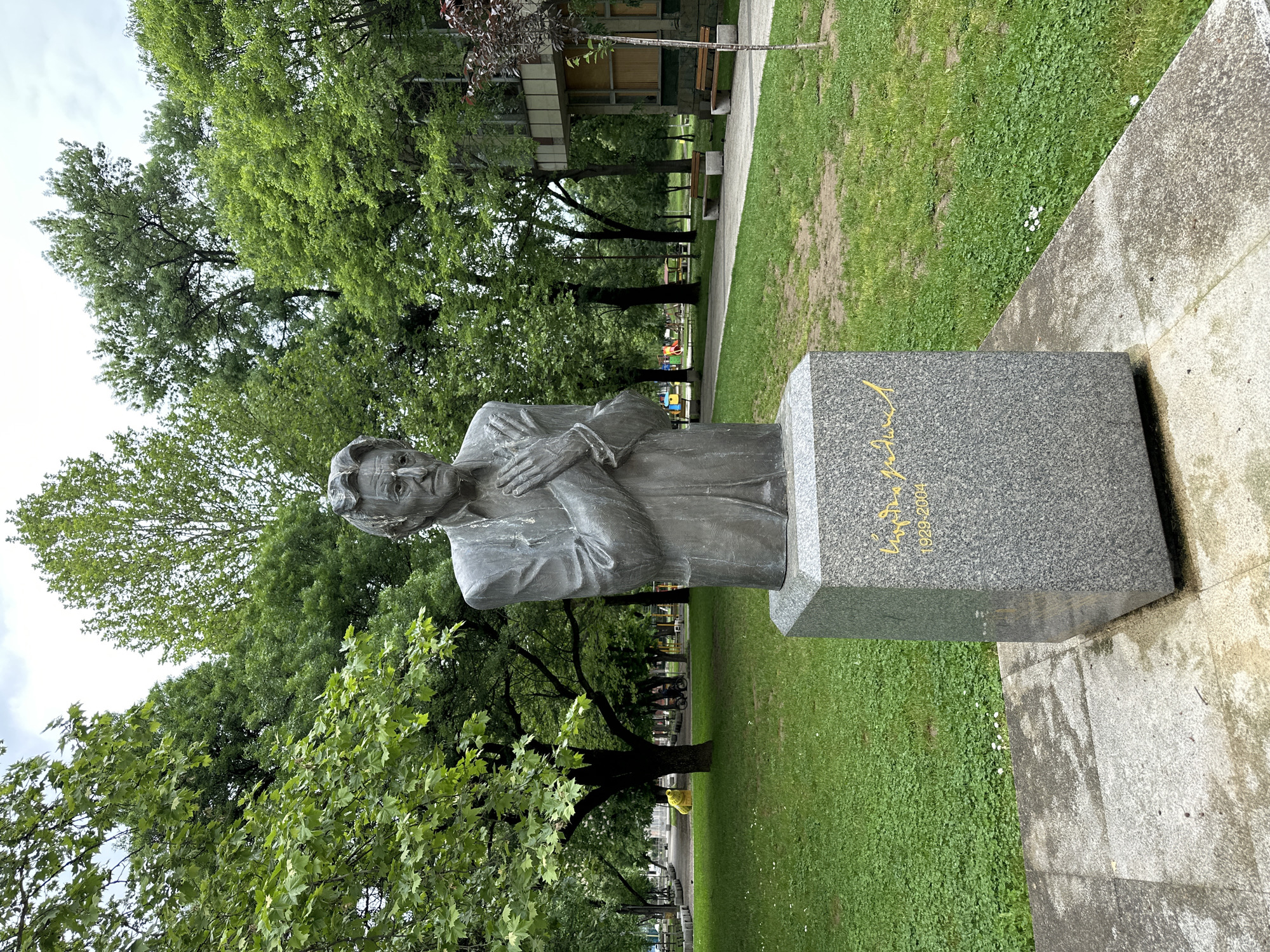 The image shows a bronze statue of a person on a granite base, standing in a green park. The statue is a bust, depicting a man with his hands clasped in front of him. The base has an inscription in gold lettering. The park has grass, trees, and benches, with a building visible in the background. The image is taken outdoors during the daytime, with overcast skies. This statue is in Montana, Bulgaria.