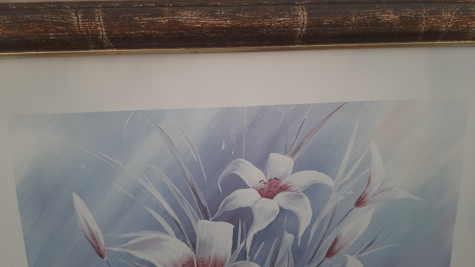 This image is a close-up of a framed painting of white lilies with pink accents against a soft blue and white background. The painting is framed with a dark, textured wood frame that has a golden inner edge. The painting appears to be an artwork rather than a photograph. The location of this artwork within the city of Desnogorsk, Russia, is not discernible from the image.