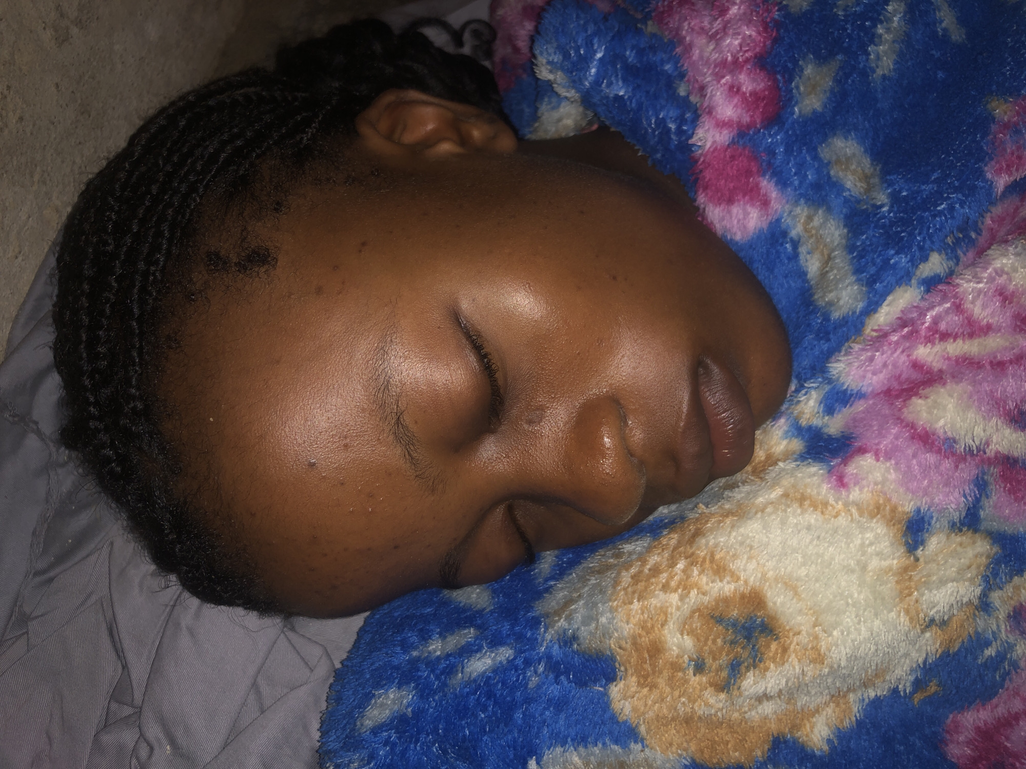 The image is a close-up, eye-level shot of a young Black girl sleeping. She is lying on her right side, facing the camera, with her eyes closed and her head resting on a blanket. The blanket has a tie-dye pattern with colors of blue, pink, and white, with some brown and yellow accents. Her hair is styled in braids. The lighting suggests an indoor setting, possibly a bedroom. The image does not contain any information that allows for the specific location in Ado, Nigeria to be identified.