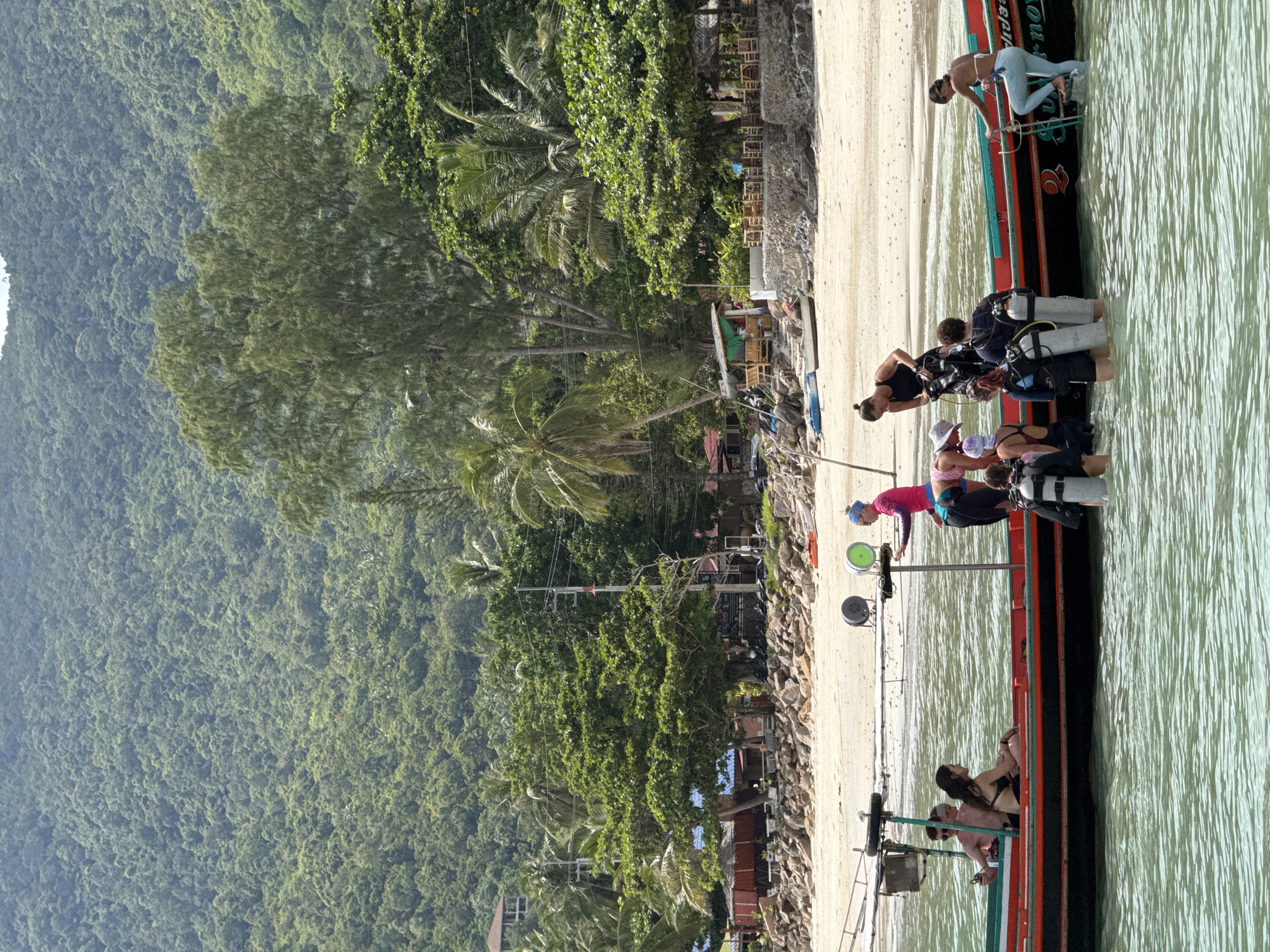 In the waters of ตำบลเกาะพะงัน, Thailand, a group of people are preparing for or returning from a diving excursion. A long, narrow boat with an orange and black hull is partially submerged in clear, greenish water. Several individuals are interacting with the boat and the water. One woman in a pink top and blue bottoms stands on the edge of the boat, pointing. Another woman in a black top and leggings is leaning over the side, assisting someone in scuba gear. Several people are already in the water wearing full scuba equipment, including tanks and masks. On the boat, two people are seated, a man and a woman, both appearing relaxed. The background features a sandy beach lined with palm trees and lush green hills that rise steeply. Buildings are visible nestled among the trees, suggesting a coastal village or resort area. The overall scene depicts a typical day for marine activities on a tropical island.