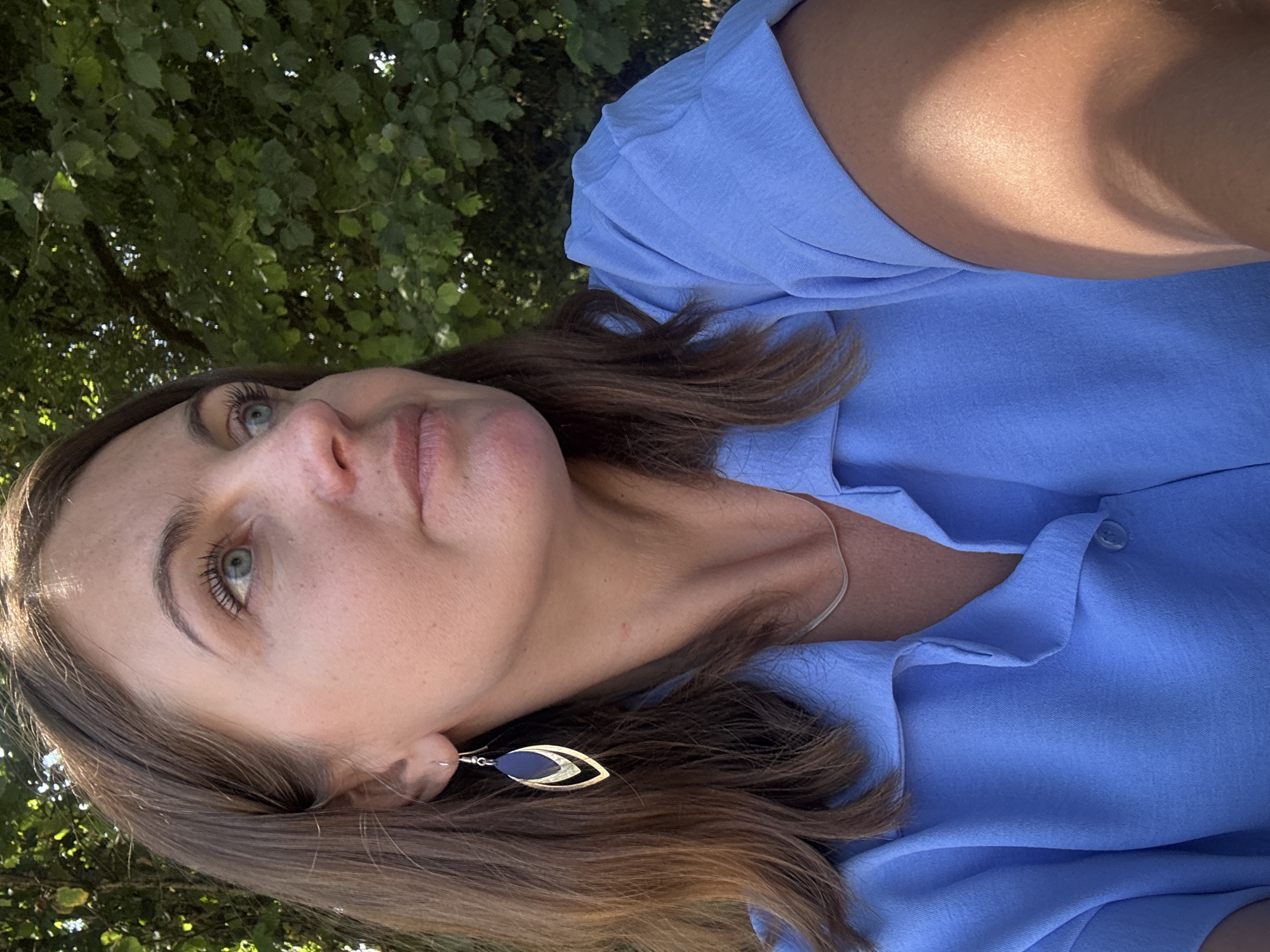 A mid-shot of a woman with brown hair and blue eyes is shown from the chest up. She is wearing a light blue button-up shirt and silver necklace. Her left ear has a dangling earring with a gold frame and a dark blue insert. She is looking to her right, with her head tilted slightly. The background consists of green foliage, suggesting an outdoor setting. The image was taken in Coteaux du Lizon, France.