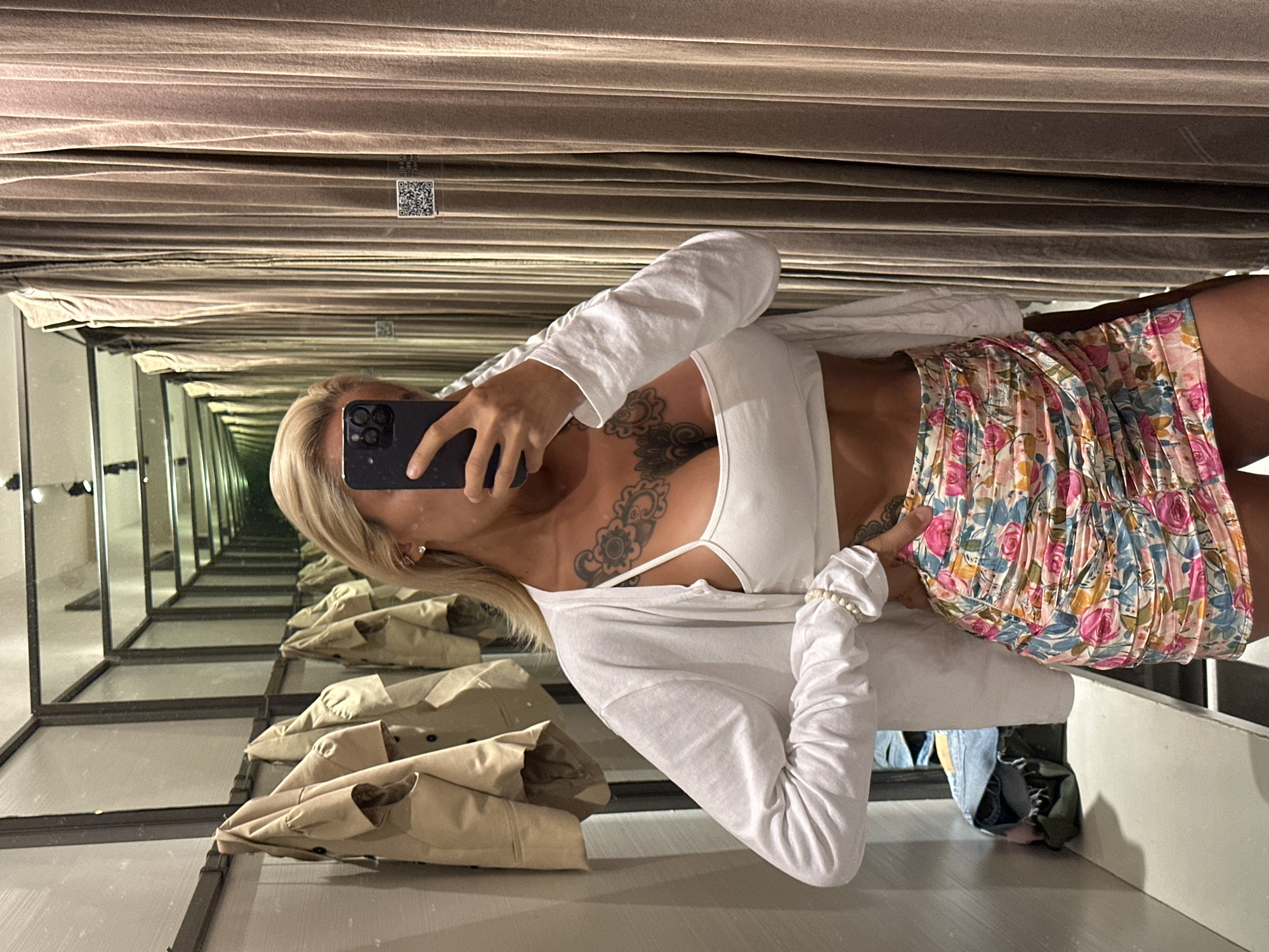 In a dimly lit fitting room, a woman with blonde hair is taking a mirror selfie. She is wearing a white crop top, a white long-sleeved cardigan, and a floral print mini skirt. She has a large tattoo on her chest and upper torso. The fitting room has beige curtains on the right side and a rack of tan jackets on the left side, with mirrors reflecting the room in the background. The woman is holding a black smartphone to take the photo. The location is not discernable from the image itself, therefore it cannot be identified as Kuta, Indonesia.